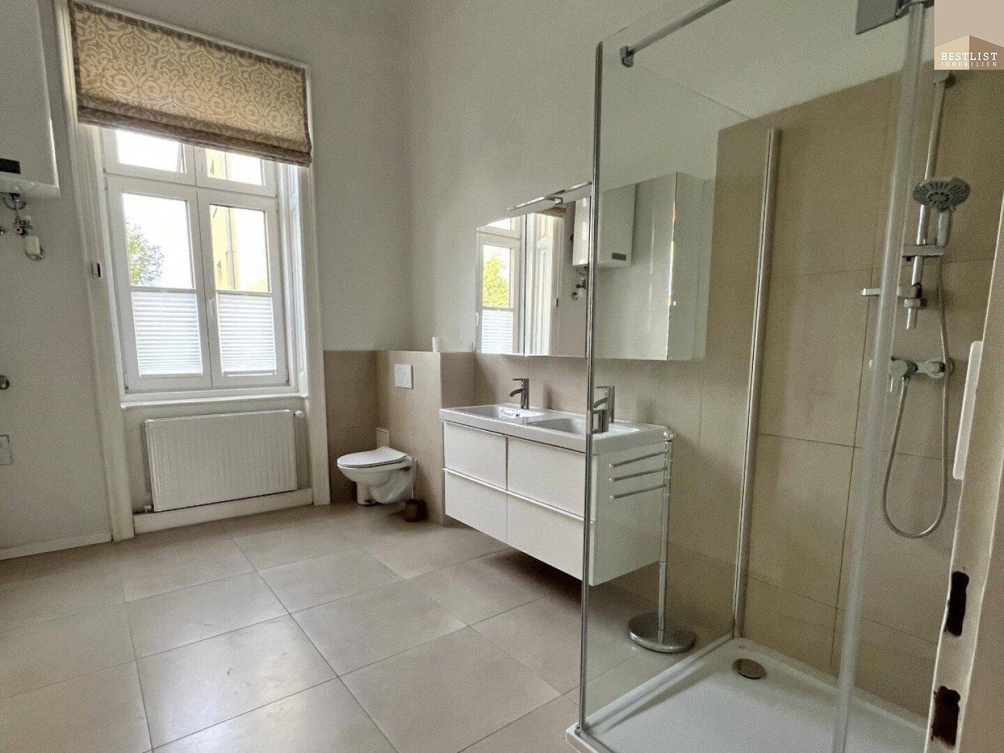 Helles Badezimmer mit Fenster, WC, Doppelwaschbecken und moderner Duschkabine, gut beleuchtet.