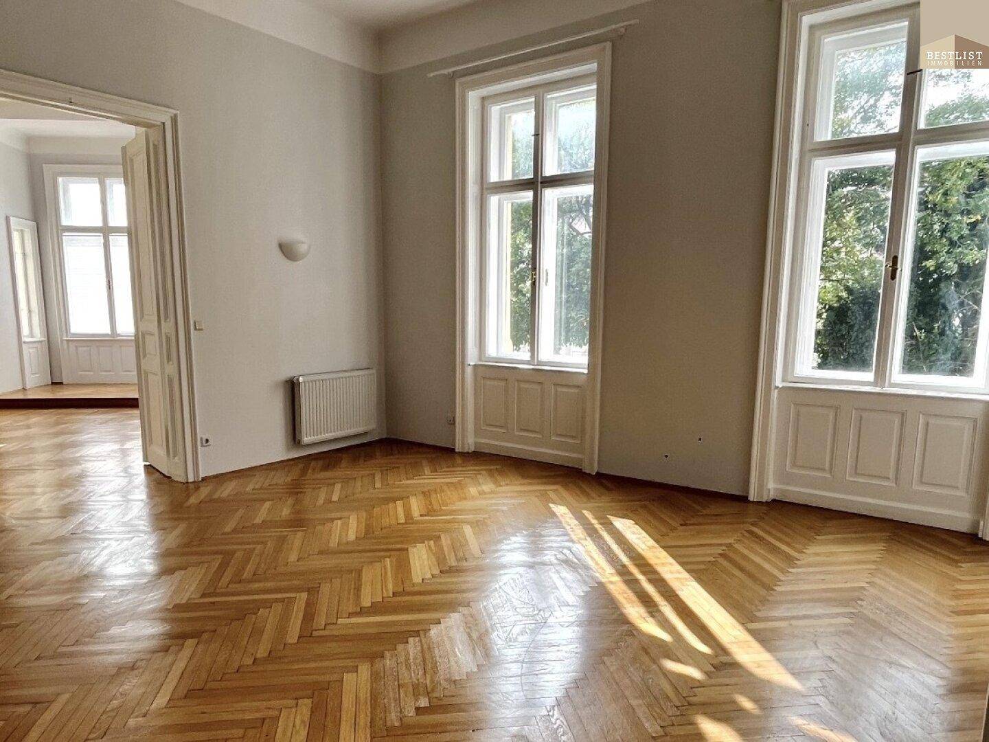 Geräumiges Wohnzimmer mit Fischgrätparkett, hohen Fenstern und viel Tageslicht, das den Raum erhellt.