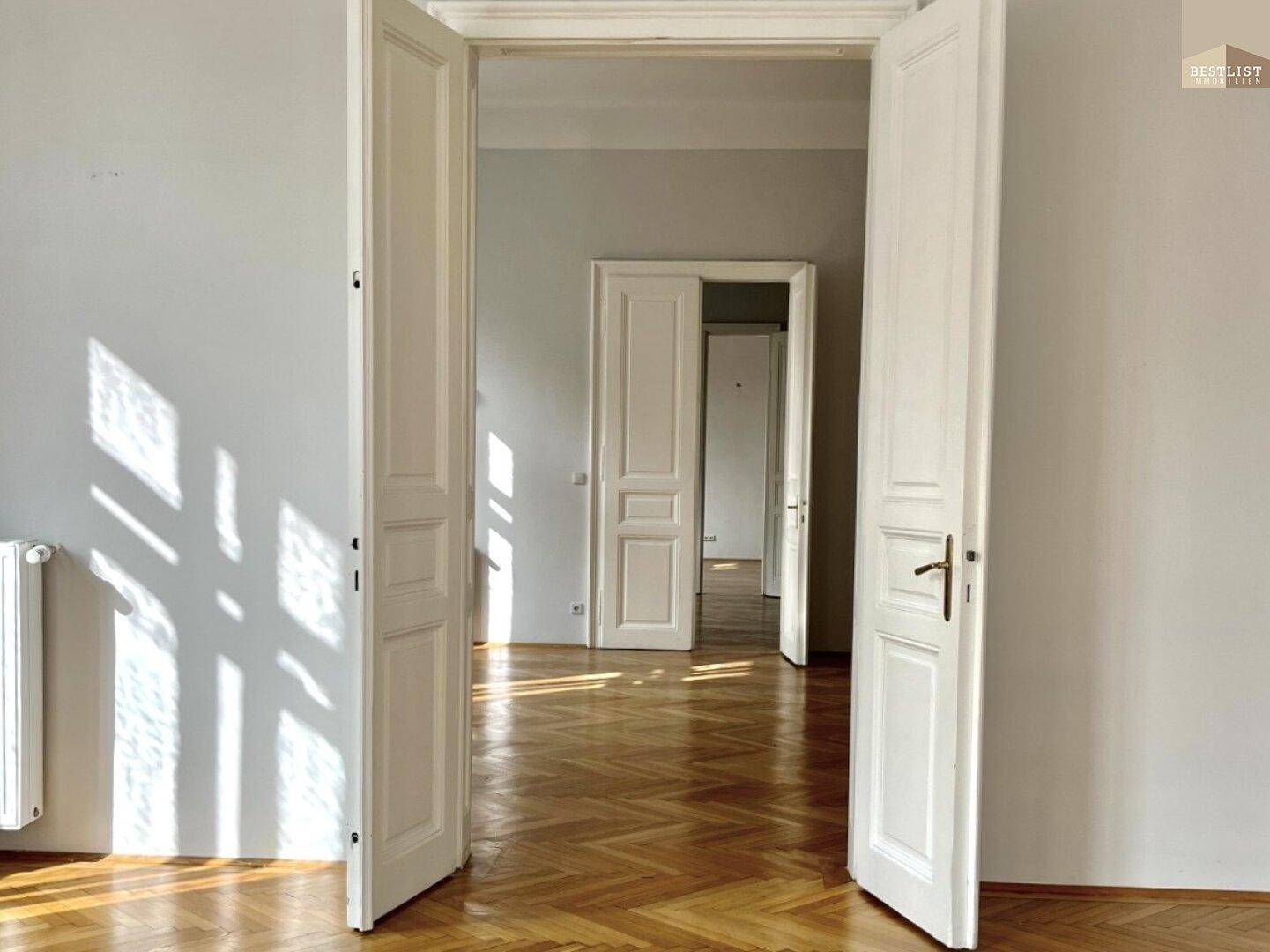 Heller Raum mit Fischgrätparkett und klassischen Flügeltüren, die weitere Zimmer in der Wohnung verbinden.