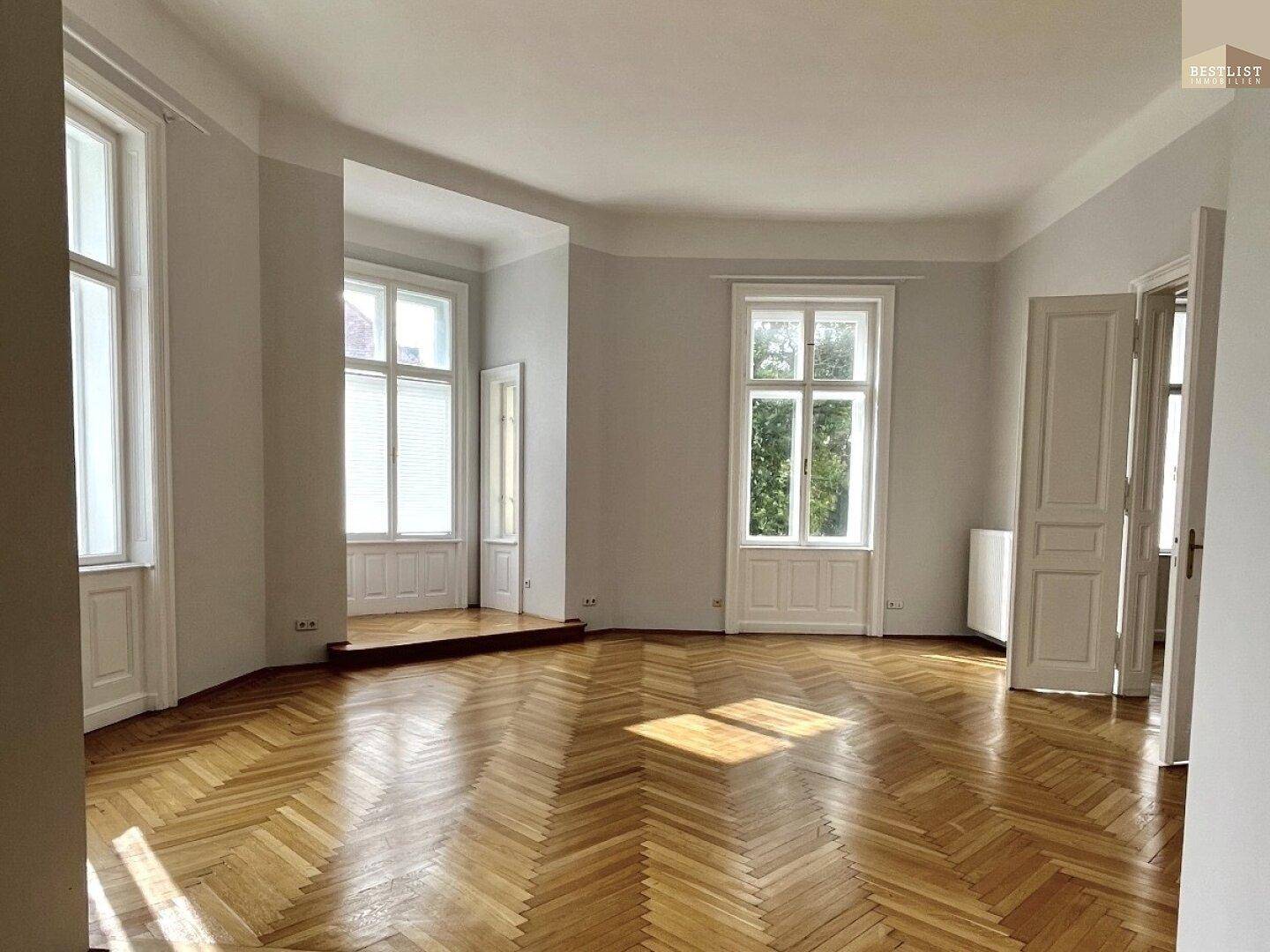 Geräumiges Zimmer mit mehreren hohen Fenstern, Fischgrätparkett und viel natürlichem Licht.