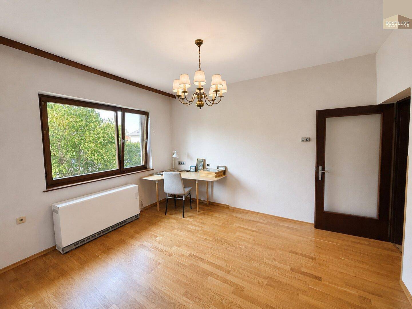 Helles Arbeitszimmer mit Holzfußboden und großem Fenster mit Blick ins Grüne.