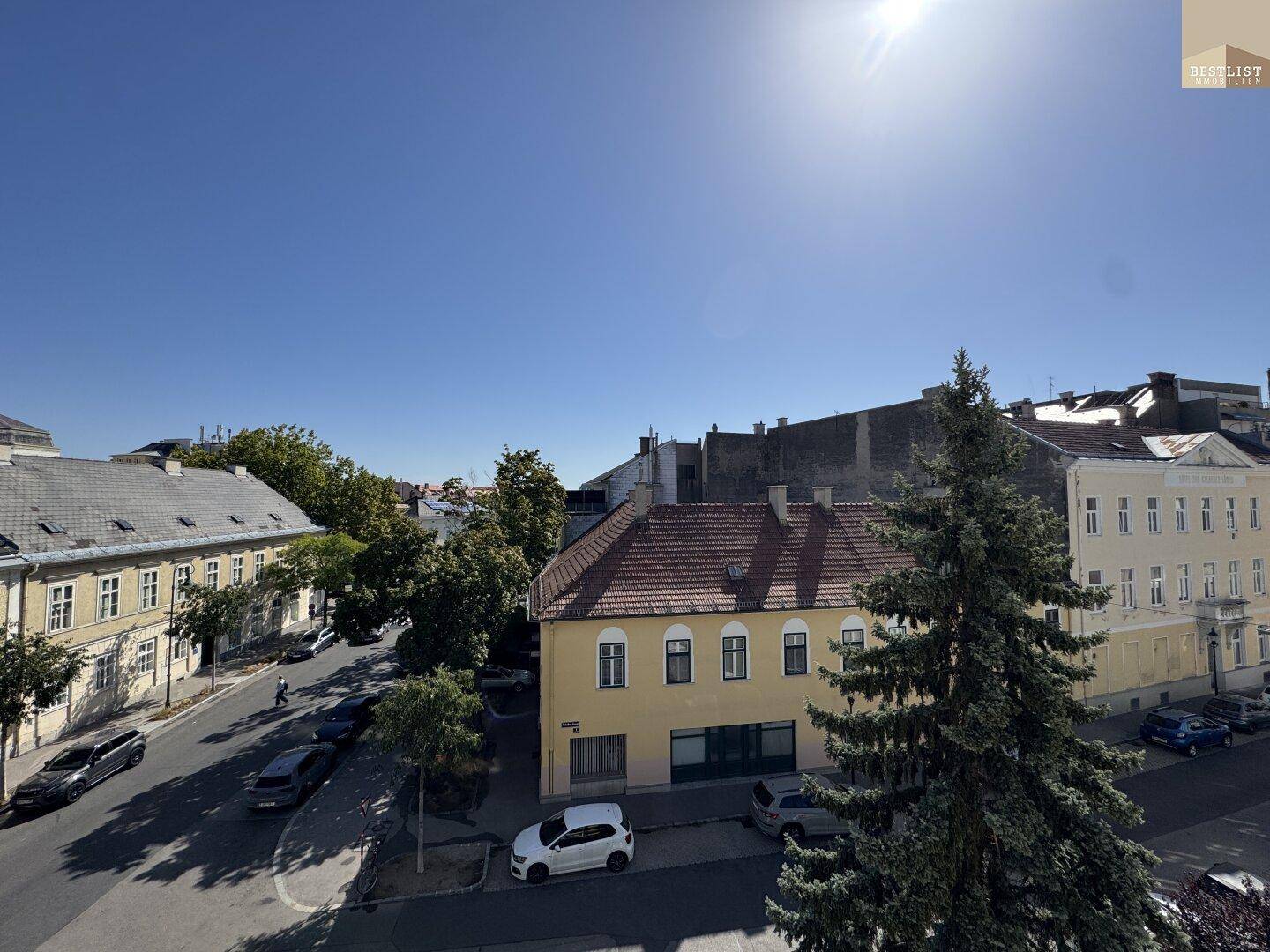 Panoramablick auf eine sonnige Straße mit historischen Gebäuden und grünen Bäumen.
