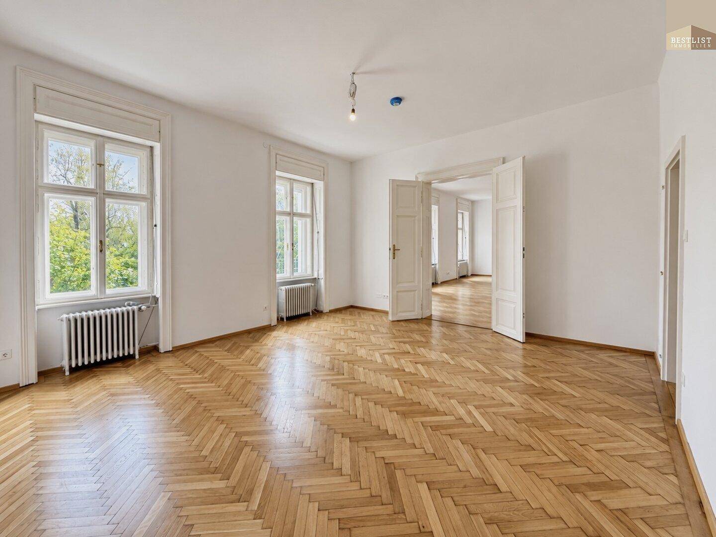 Eleganter Wohnraum mit Fischgrätparkett, hohen Fenstern und Flügeltüren zu angrenzenden Zimmern.
