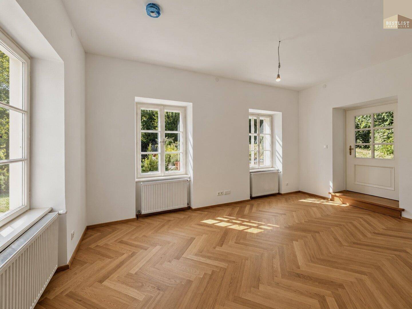 Lichtdurchfluteter Raum mit Fischgrätparkett, Blick ins Grüne und klassischer Heizkörperausstattung.