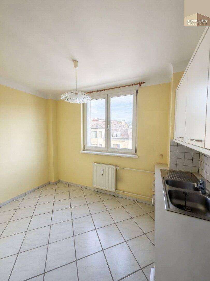 Heller Küchenbereich mit Fliesenboden, Fenster und Heizkörper, bereit für eine Essecke.