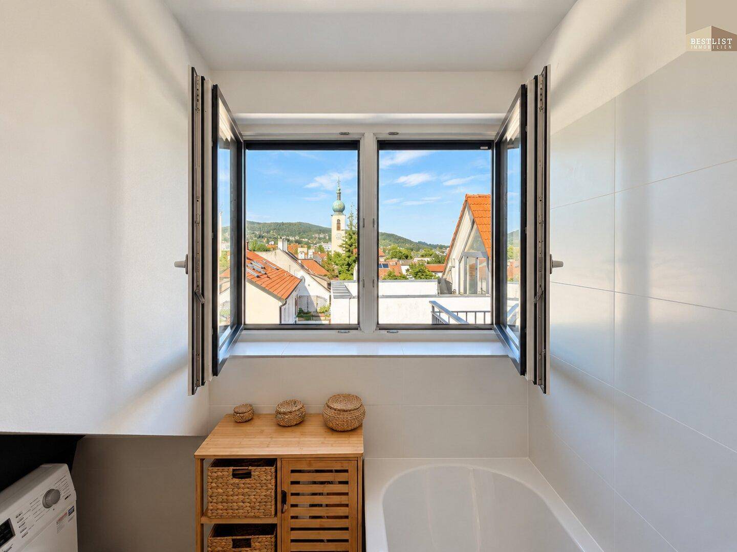 Helles Badezimmer mit Fenster, Badewanne und Ausblick auf die Stadt und Berge.