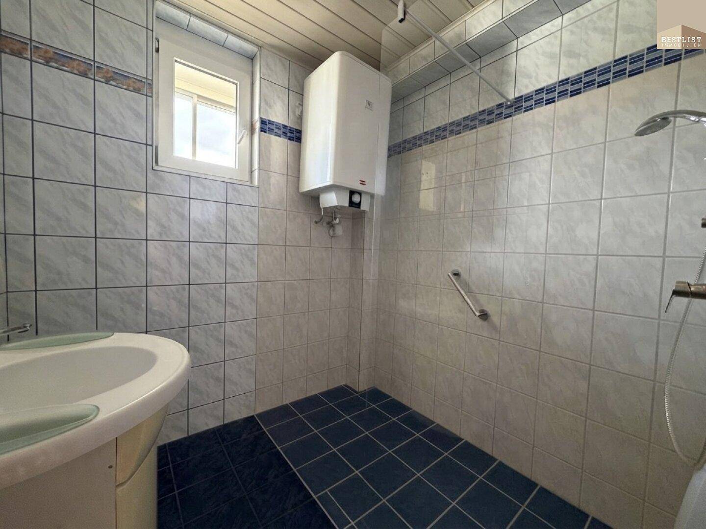Badezimmer mit blauen und weißen Fliesen, Dusche, Fenster und praktischen Haltegriffen.