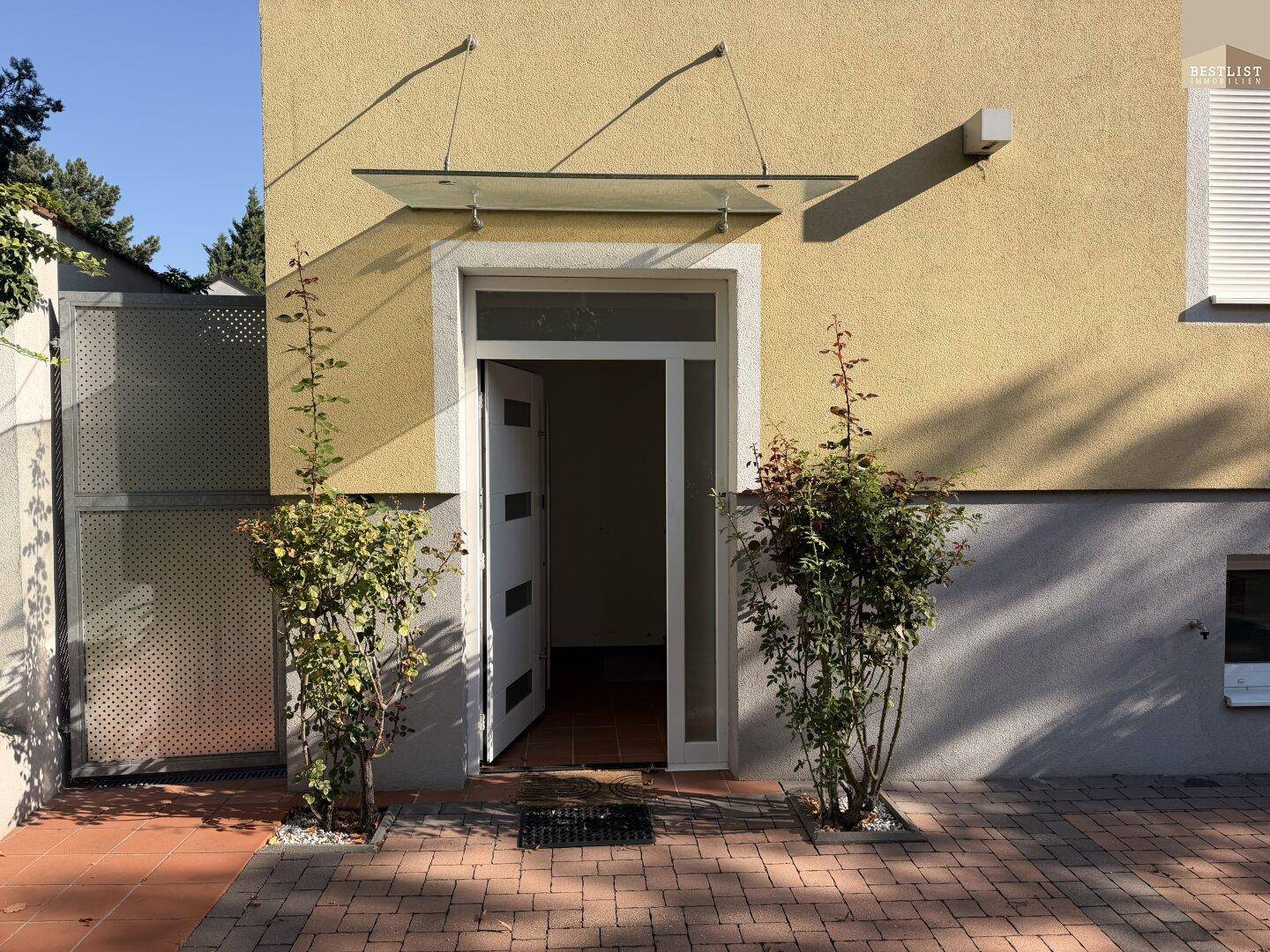 Eingangsbereich des Hauses mit einer Glastür und gepflastertem Vorplatz unter klarem Himmel.