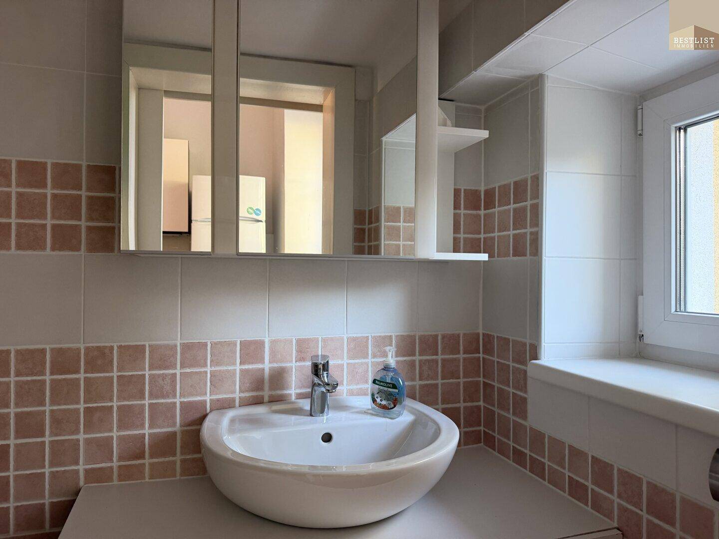 Badezimmer mit Waschbecken, Spiegelschrank und beigefarbenen Fliesen, gut beleuchtet durch ein Fenster.