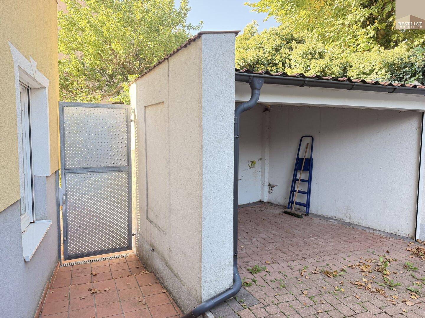 Einfahrt zu einem überdachten Stellplatz mit einem Metalltor und gepflastertem Boden.