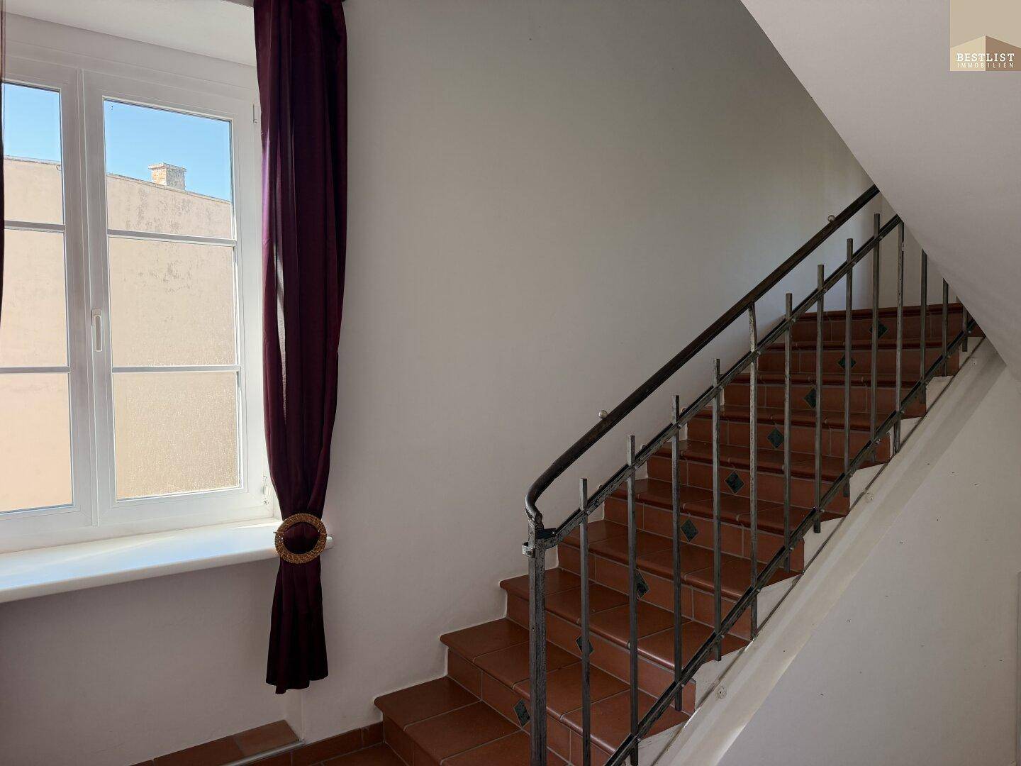 Treppenhaus mit Terrakottafliesen und schmiedeeisernem Geländer, beleuchtet durch ein Fenster.