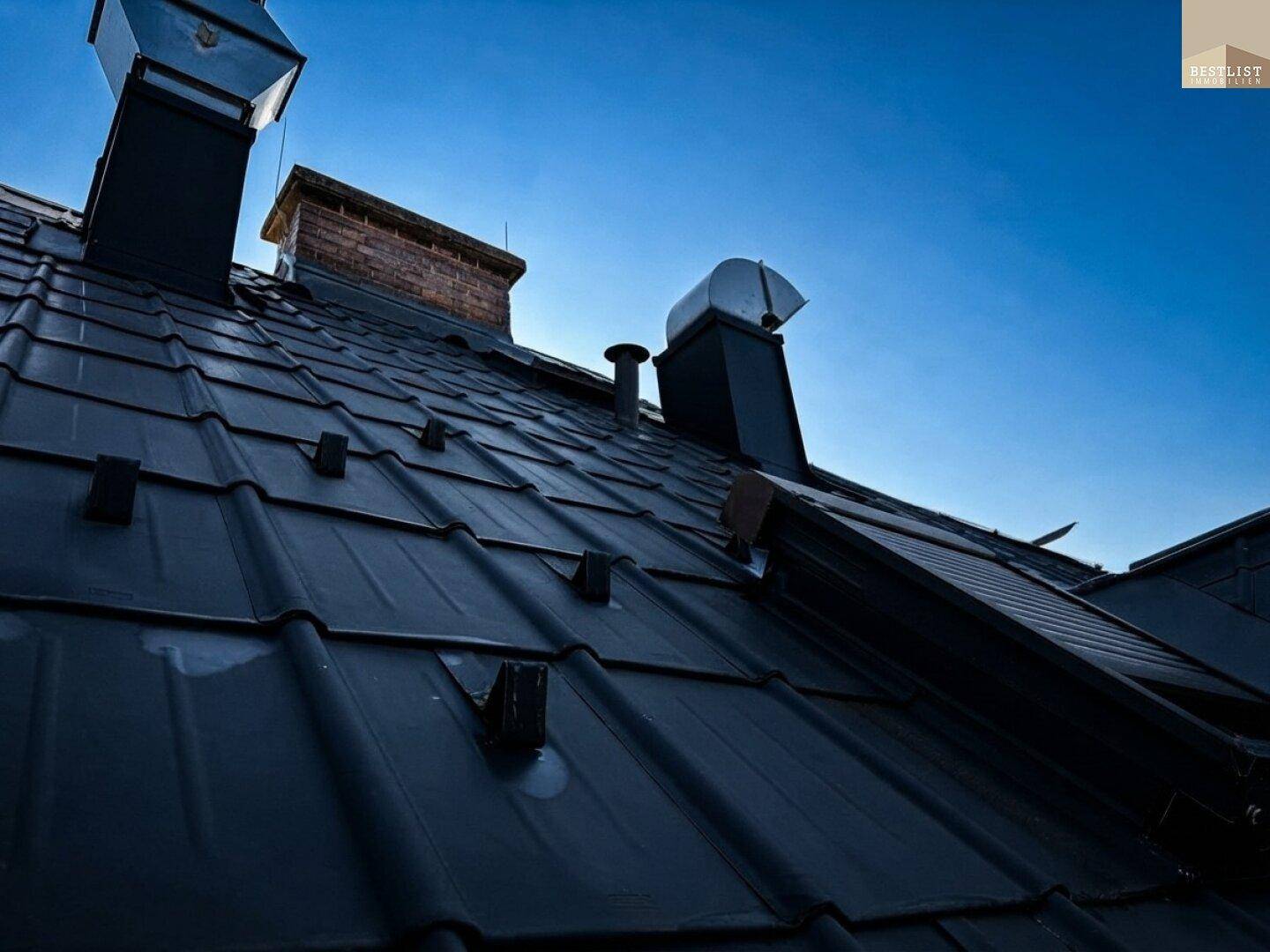 Schwarzes Ziegeldach mit Schornsteinen unter klarem blauem Himmel.