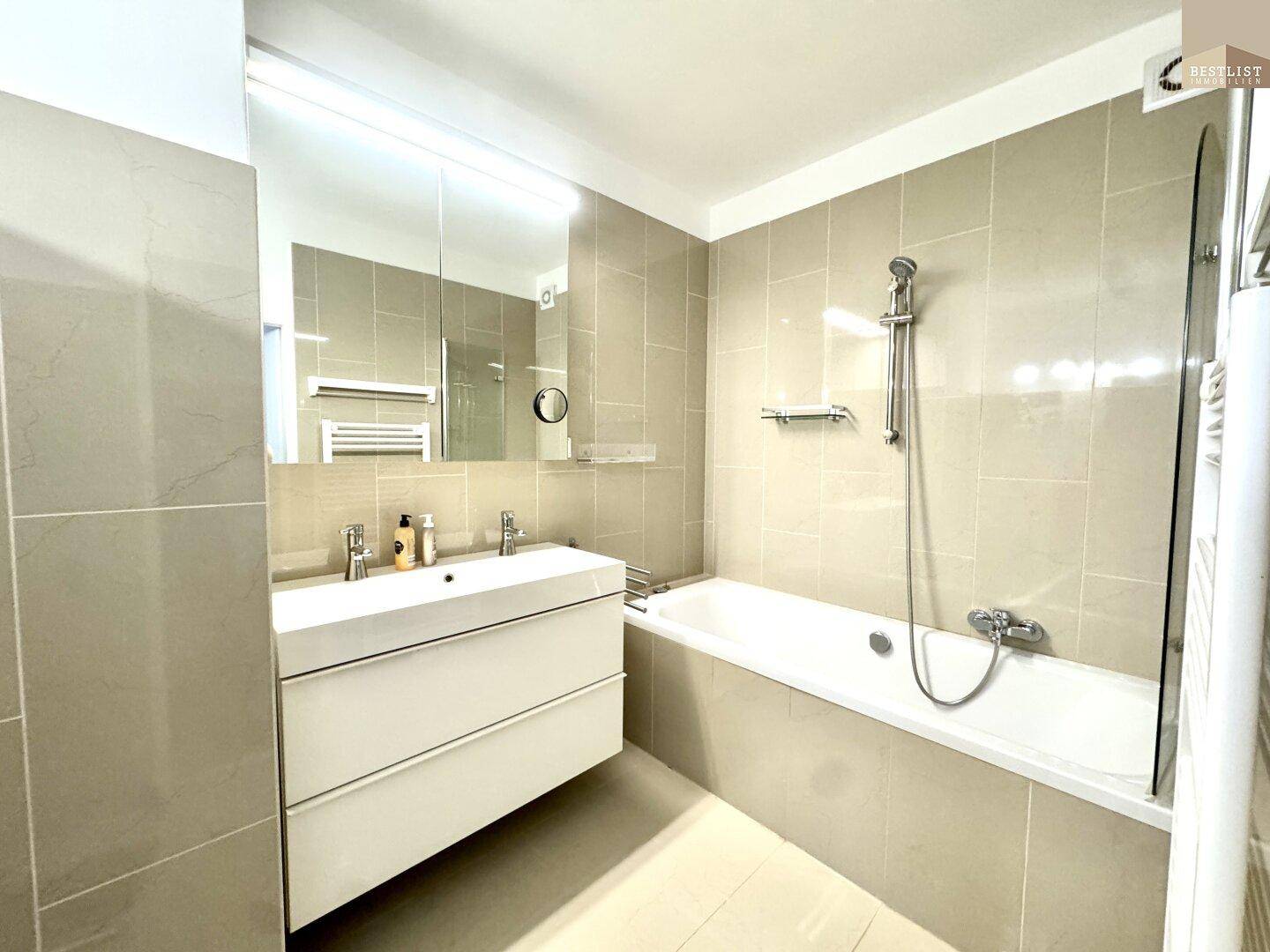 Modernes Badezimmer mit Badewanne, Doppelwaschbecken und hellen Fliesen an Wänden und Boden.