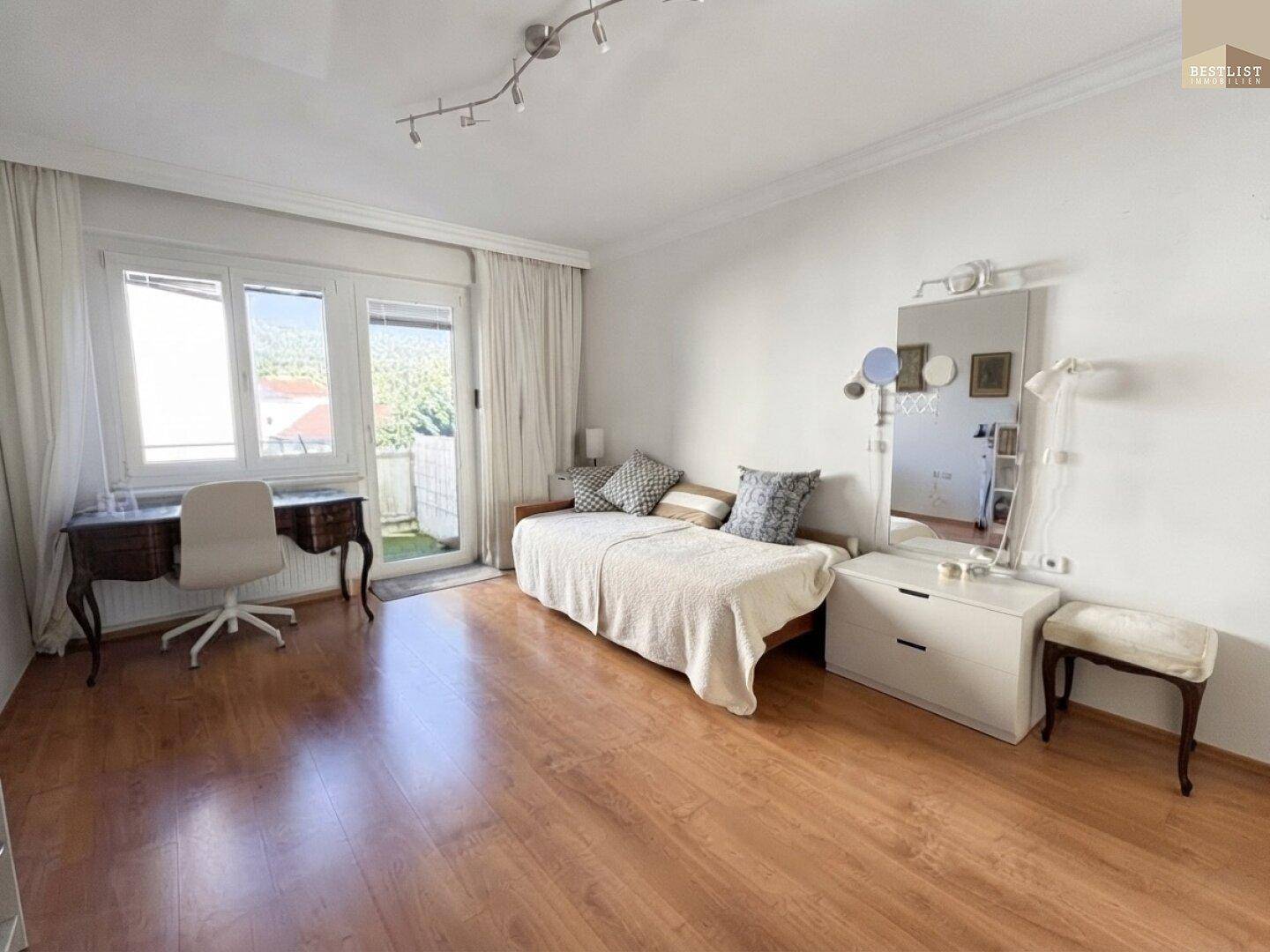 Schlafzimmer mit Holzboden, Schreibtisch und direktem Zugang zu einem sonnigen Balkon.