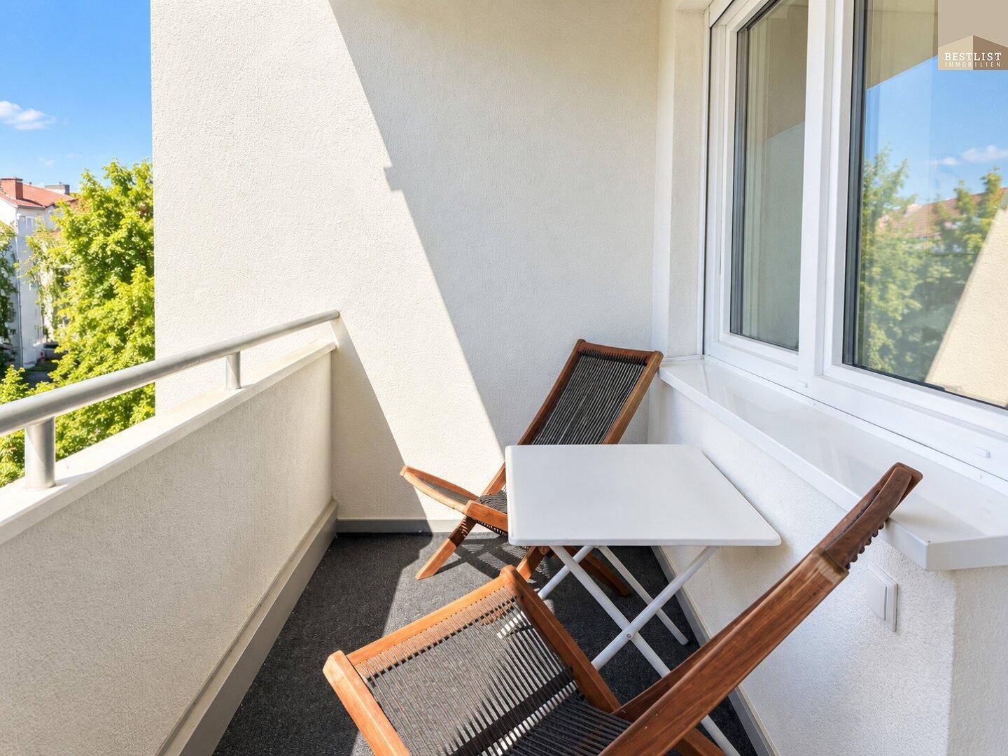 Kleiner, sonniger Balkon mit Sitzgelegenheiten und Blick auf die umliegenden Bäume.