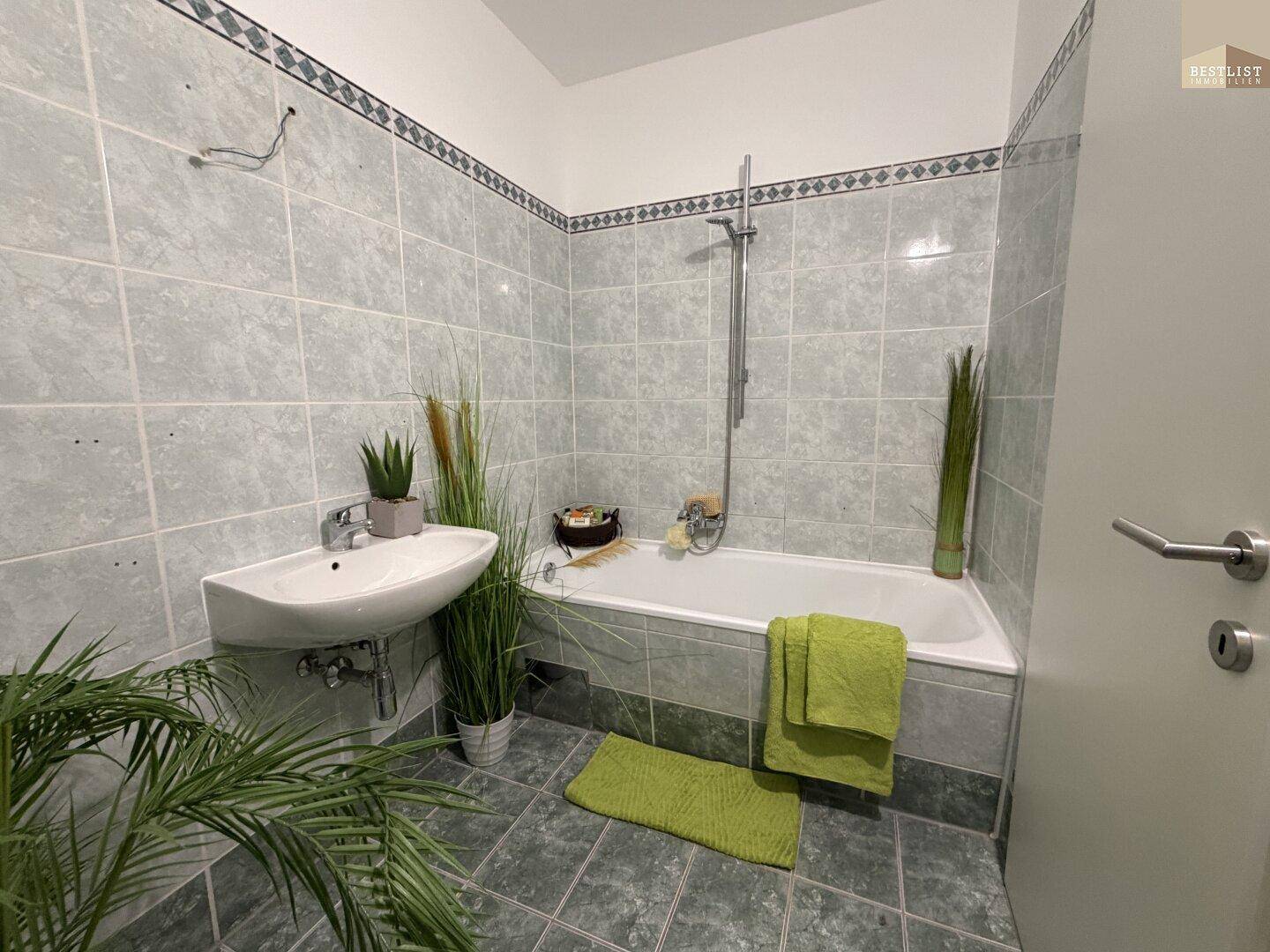 Badezimmer mit Badewanne, Waschbecken und hellgrauen Fliesen, dekoriert mit Pflanzen.