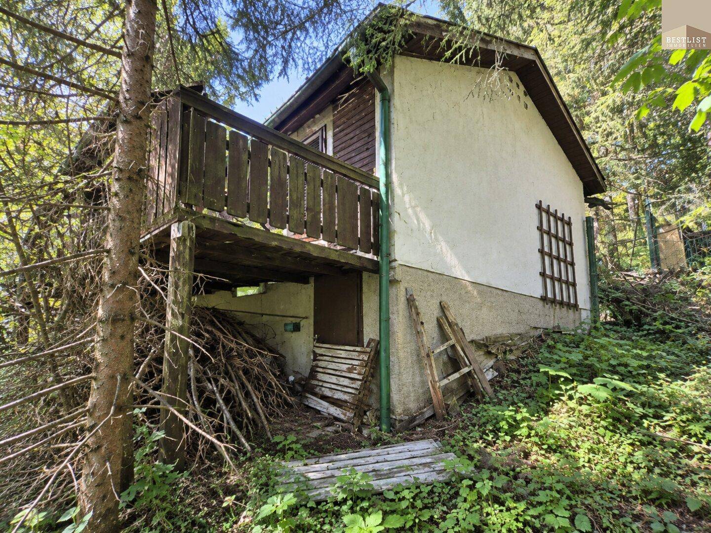 Äußere Ansicht eines Hauses im Wald mit baufälligem Holzbalkon und viel Grün in der Umgebung.