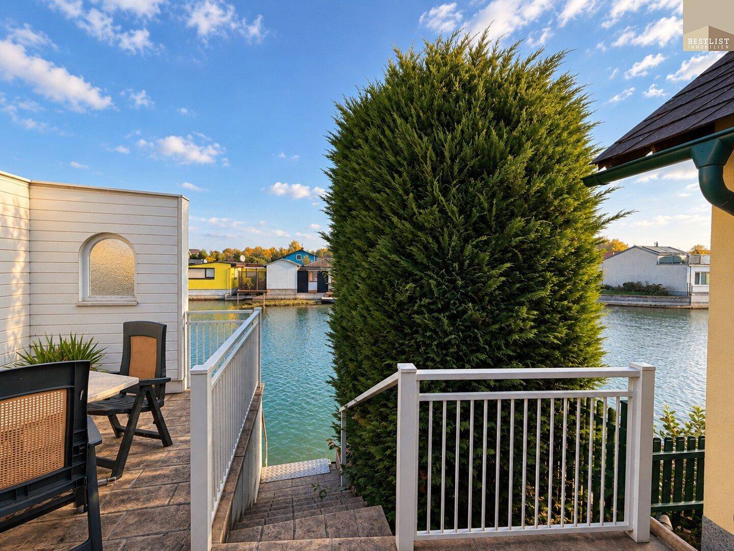 Terrasse am Wasser mit Sitzgelegenheiten und Blick auf den See oder Kanal unter blauem Himmel.