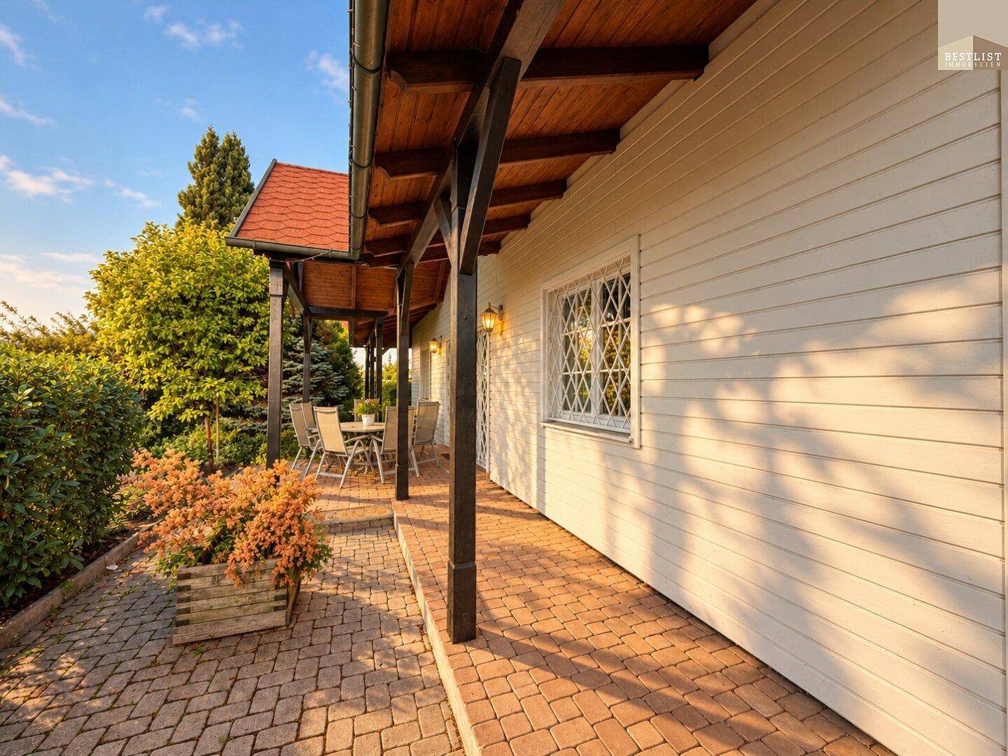 Langer überdachter Terrassenbereich mit Pflastersteinen und Blumenkübeln entlang der Hausfassade.