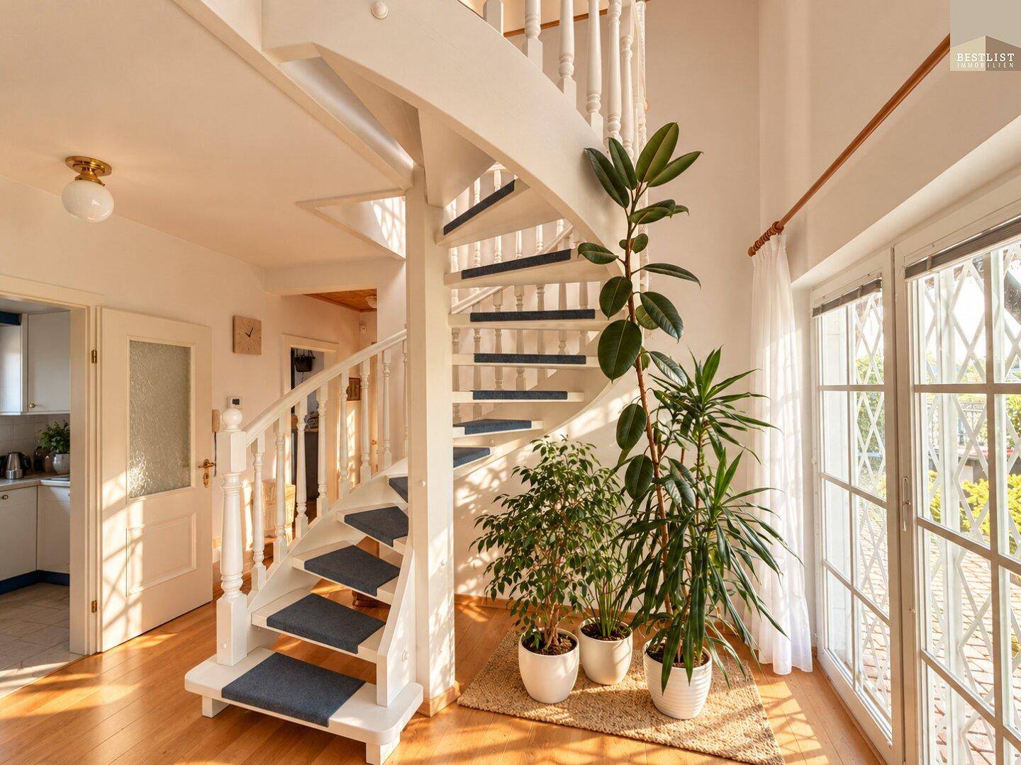 Heller Eingangsbereich mit eleganter Wendeltreppe, Holzfußboden und großen Fenstern, die viel Licht hereinlassen.