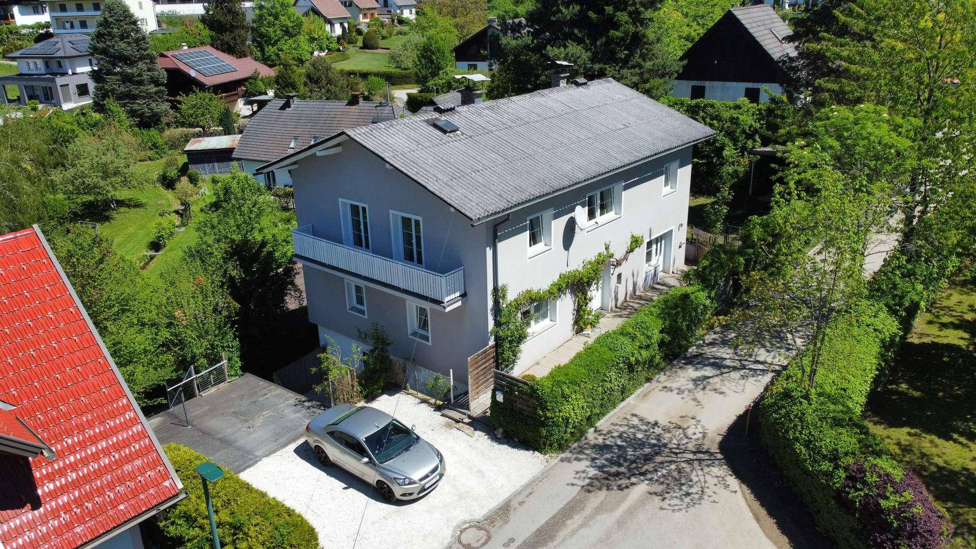 Ansicht eines grauen Wohnhauses mit Balkon, umgeben von Bäumen und einer Zufahrt mit geparktem Auto.