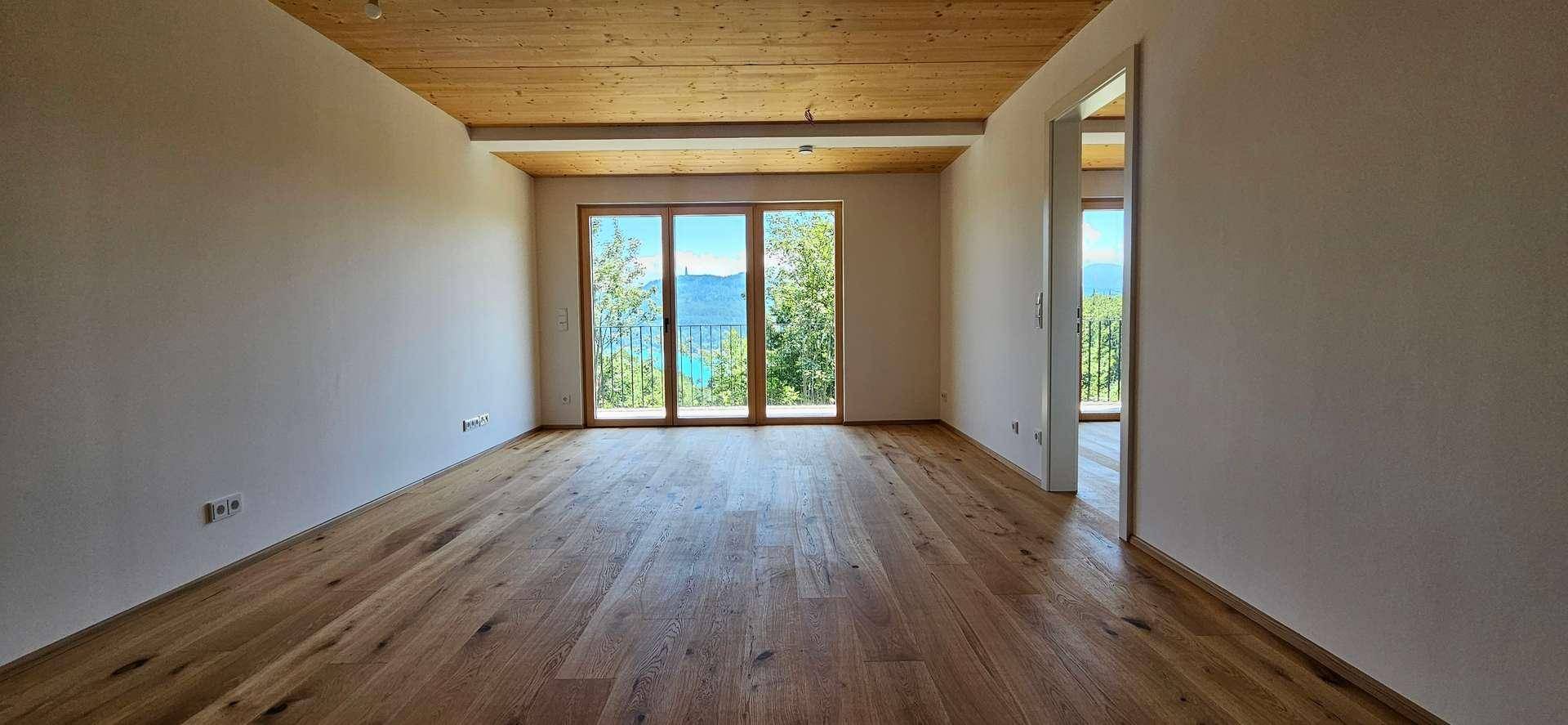Heller Raum mit Holzboden und Holzdecke, großen Fenstern und Balkonzugang mit Seeblick.