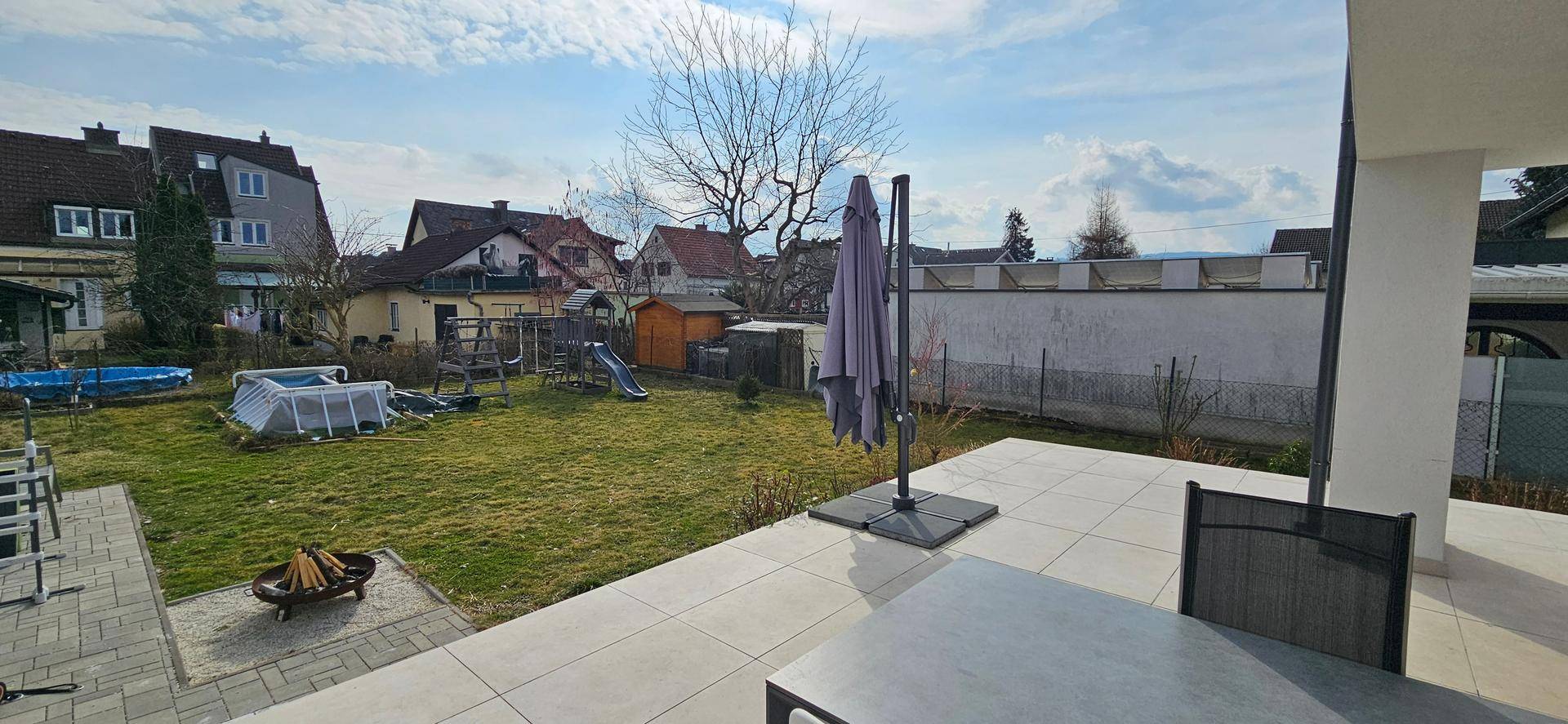 Sonniger Gartenbereich mit großer Terrasse, Gartenmöbeln, Feuerstelle und Spielplatz.