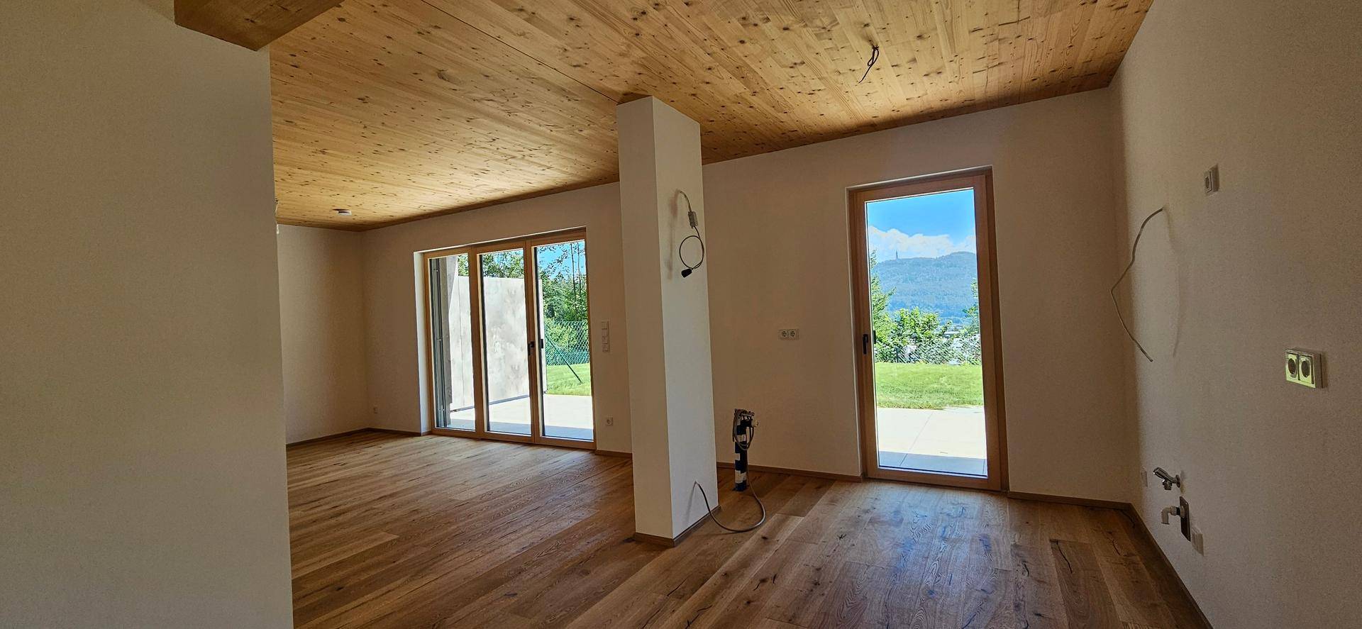 Leerer Raum mit Holzboden und Holzdecke, großen Fenstern und weitem Blick ins Grüne.