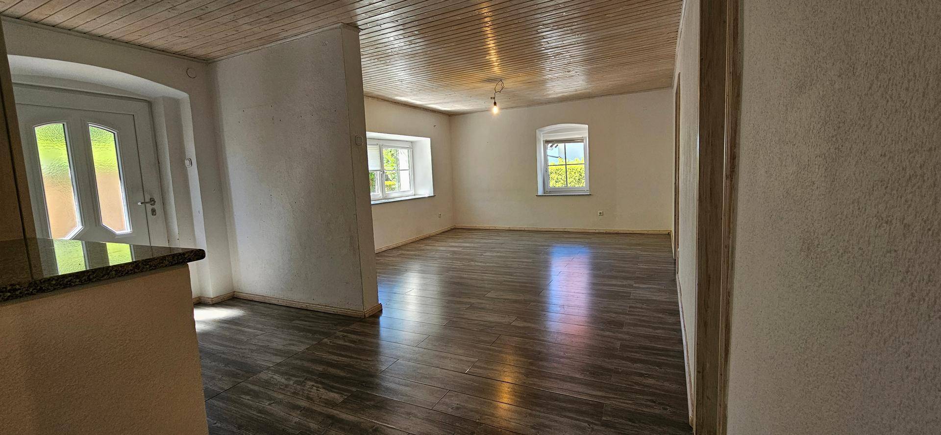Heller Wohnbereich mit Holzboden, Holzdecke und zwei Fenstern, die viel Tageslicht hereinlassen.
