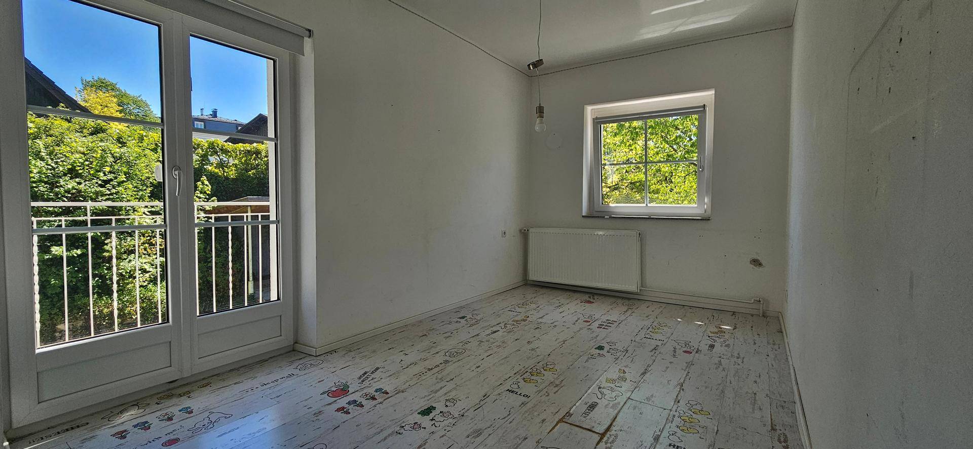 Helles Schlafzimmer mit Fenster, Heizung und Zugang zu einem kleinen Balkon mit Blick ins Grüne.