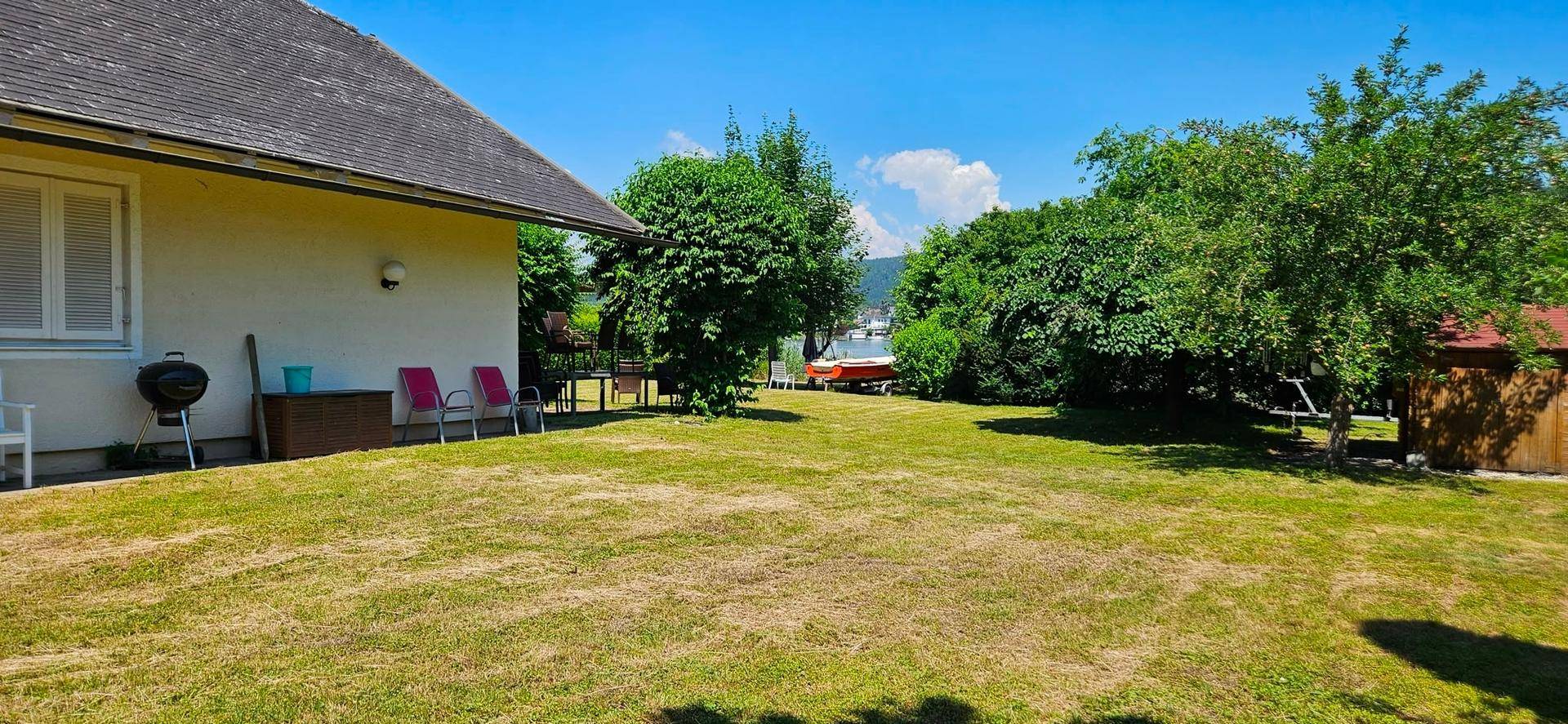 Seitlicher Blick auf das Haus mit großem Rasenbereich, Grill und Gartenmöbeln unter blauem Himmel.