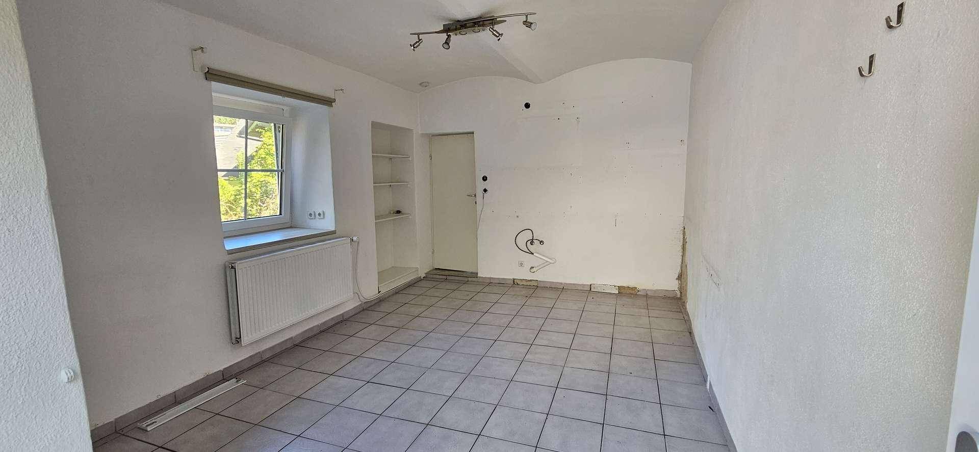Leerer Raum mit Fliesenboden, einem Fenster und Anschlussmöglichkeiten an der Wand, bereit zur individuellen Gestaltung.