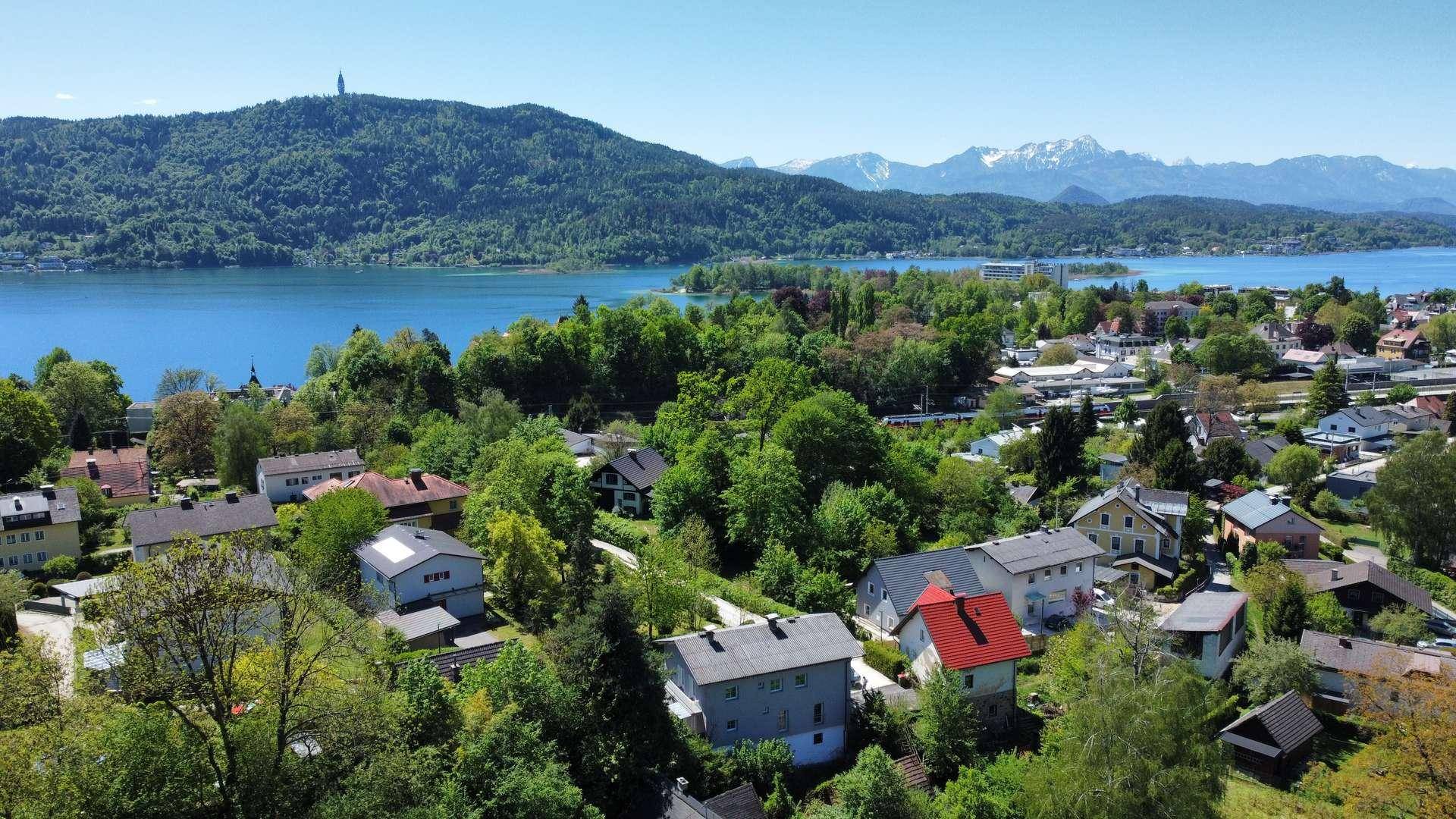 Weitläufige Luftaufnahme einer Wohngegend mit See, bewaldeten Hügeln und schneebedeckten Bergen am Horizont.