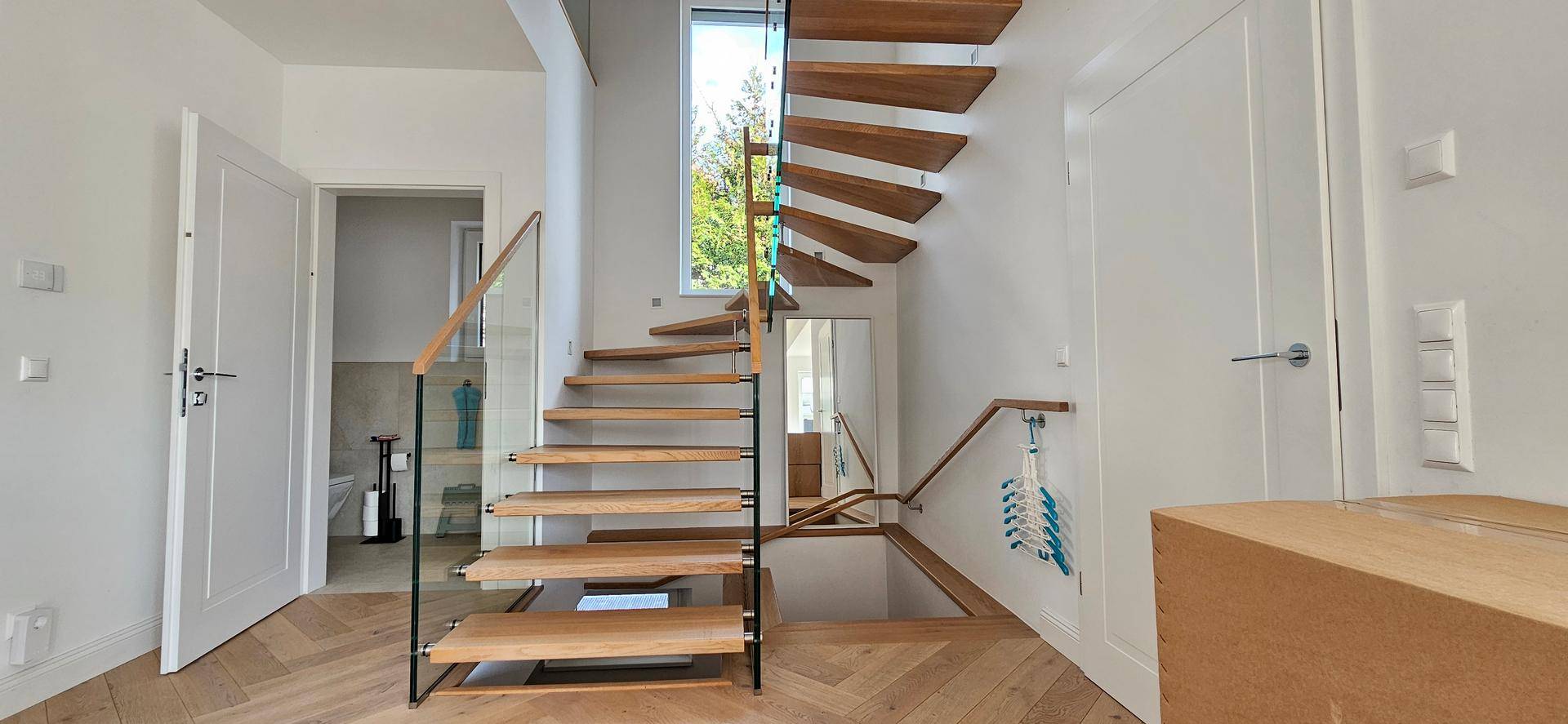 Moderner Flur mit offener Holztreppe, Glasgeländer und hellem Fischgrätparkettboden.