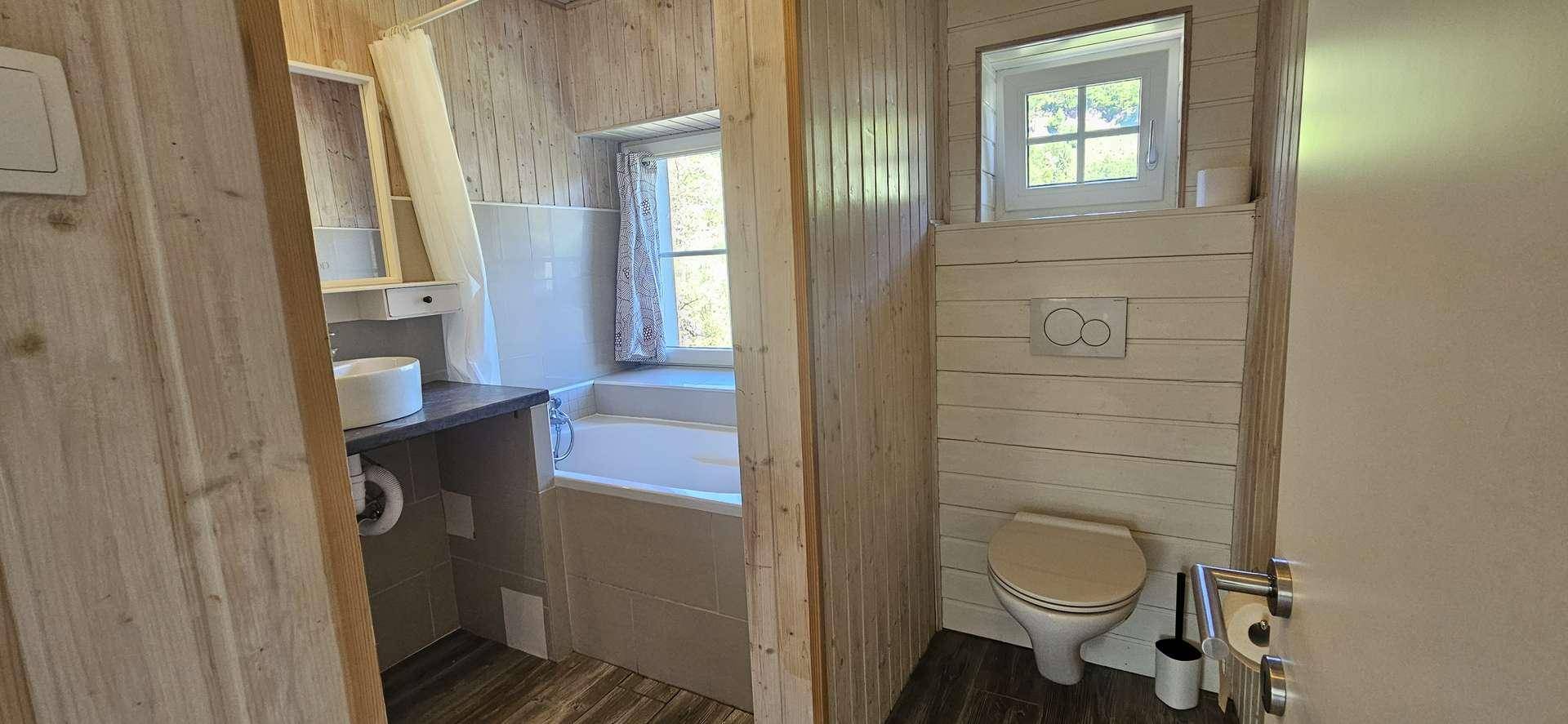 Rustikales Badezimmer mit Holzverkleidung, Badewanne, WC und einem kleinen Fenster für natürliches Licht.