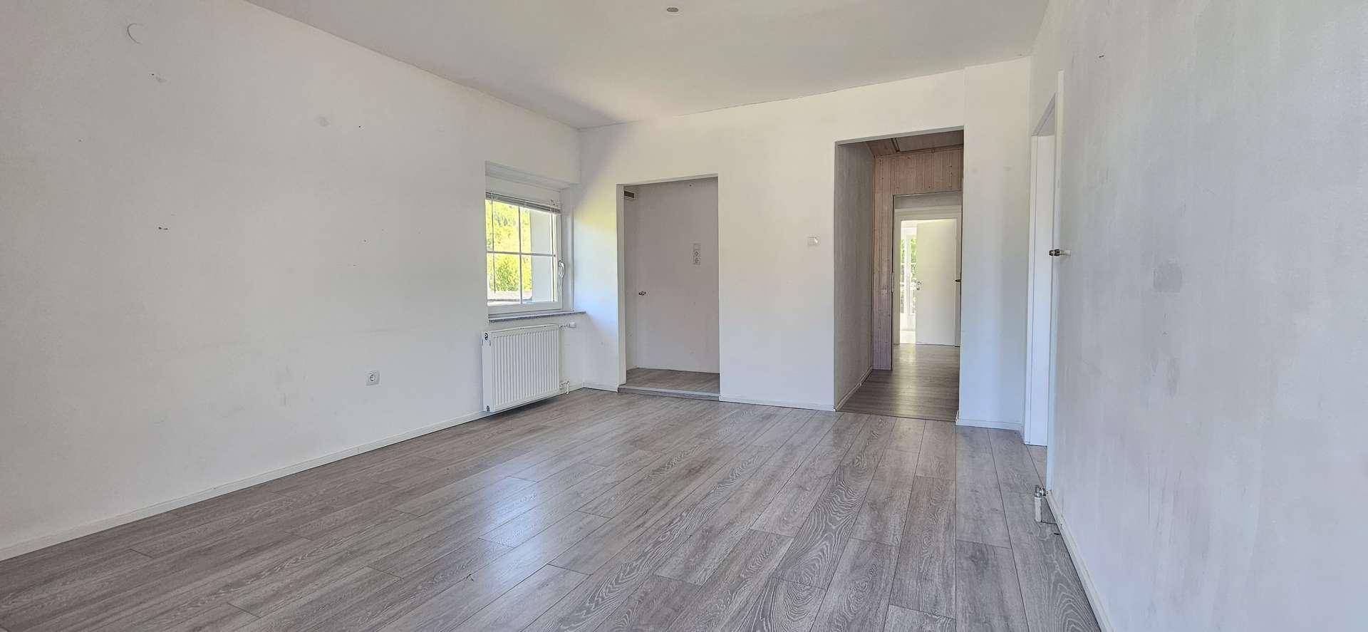 Heller Wohnraum mit Laminatboden, weißen Wänden und einem Fenster, das viel Tageslicht hereinlässt.