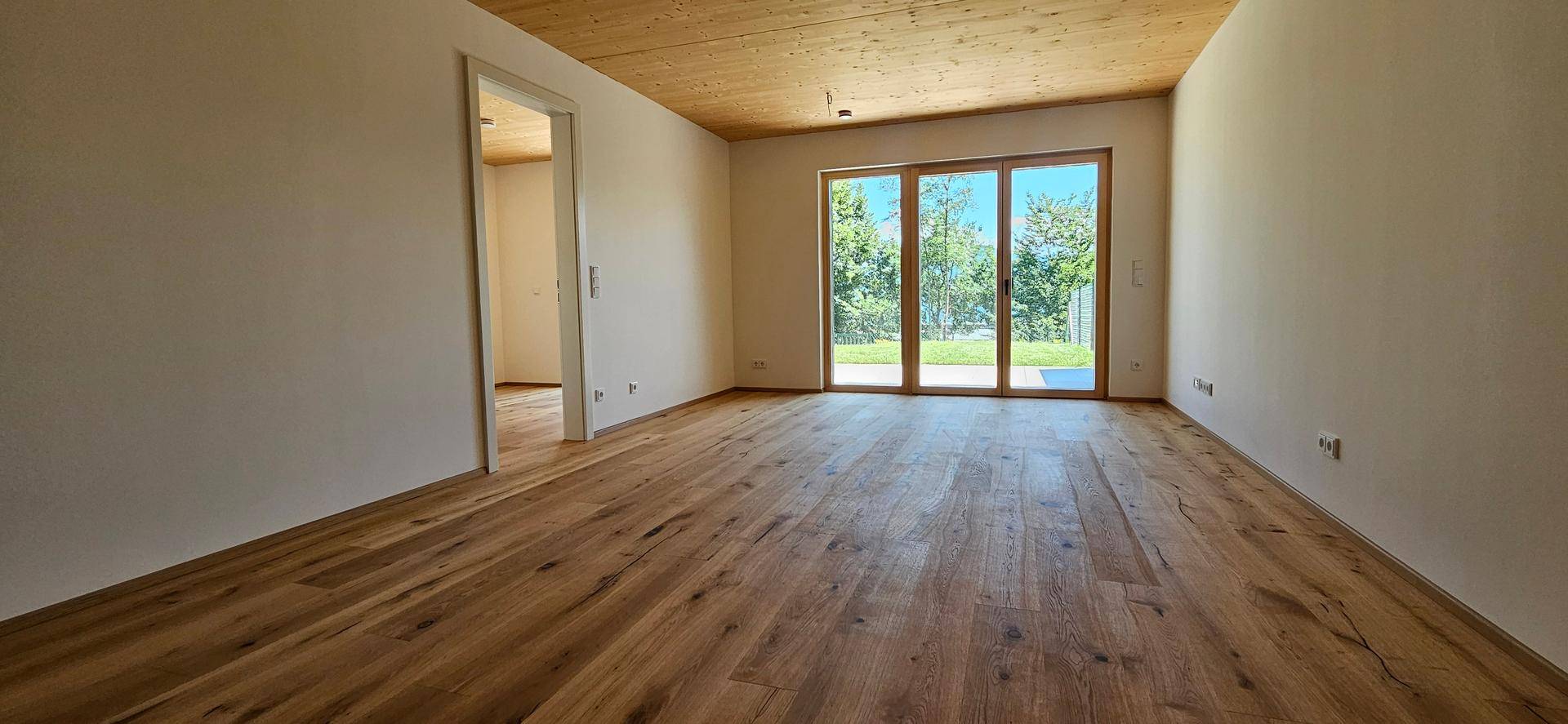 Heller, leerer Wohnraum mit Holzboden, Holzdecke und großen Fenstern zum Garten.