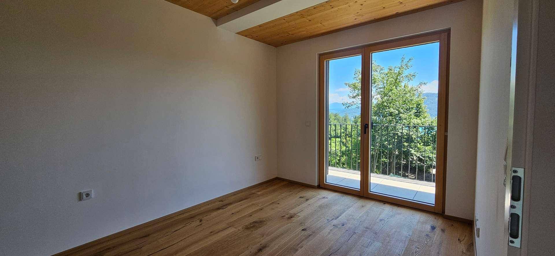 Leeres Schlafzimmer mit Holzboden, Holzdecke und großen Fenstern mit Blick ins Grüne.