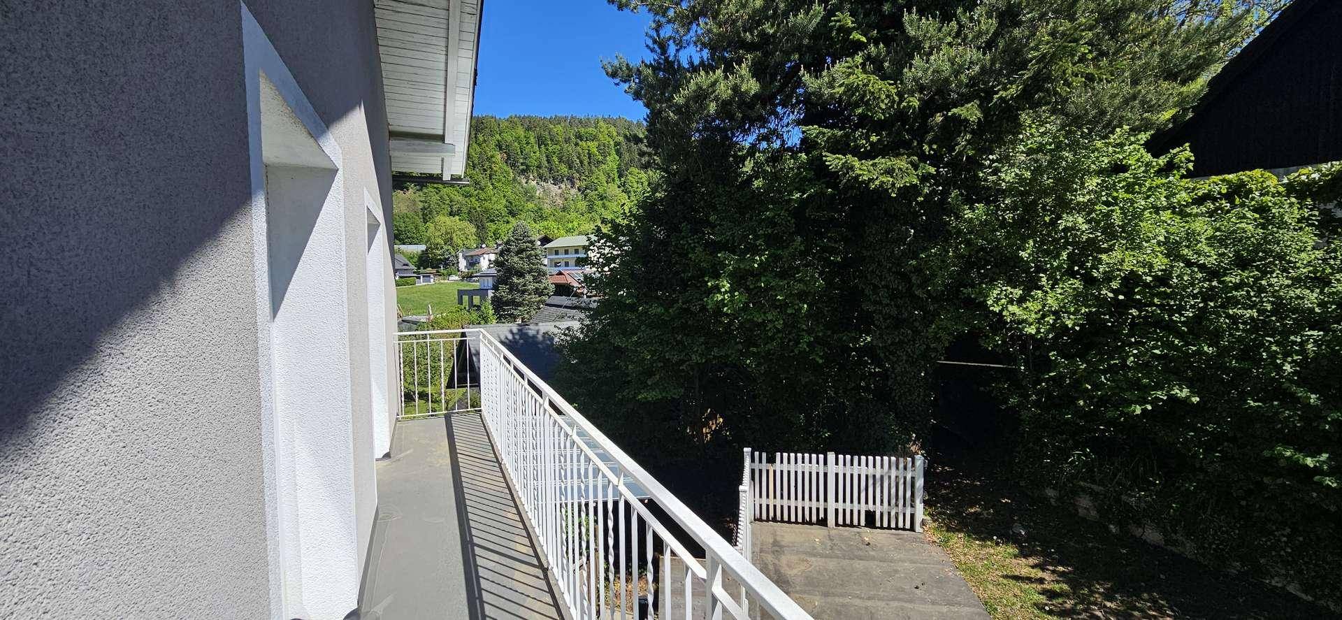 Heller Balkon mit weißem Geländer, umgeben von üppigem Grün und Bäumen.