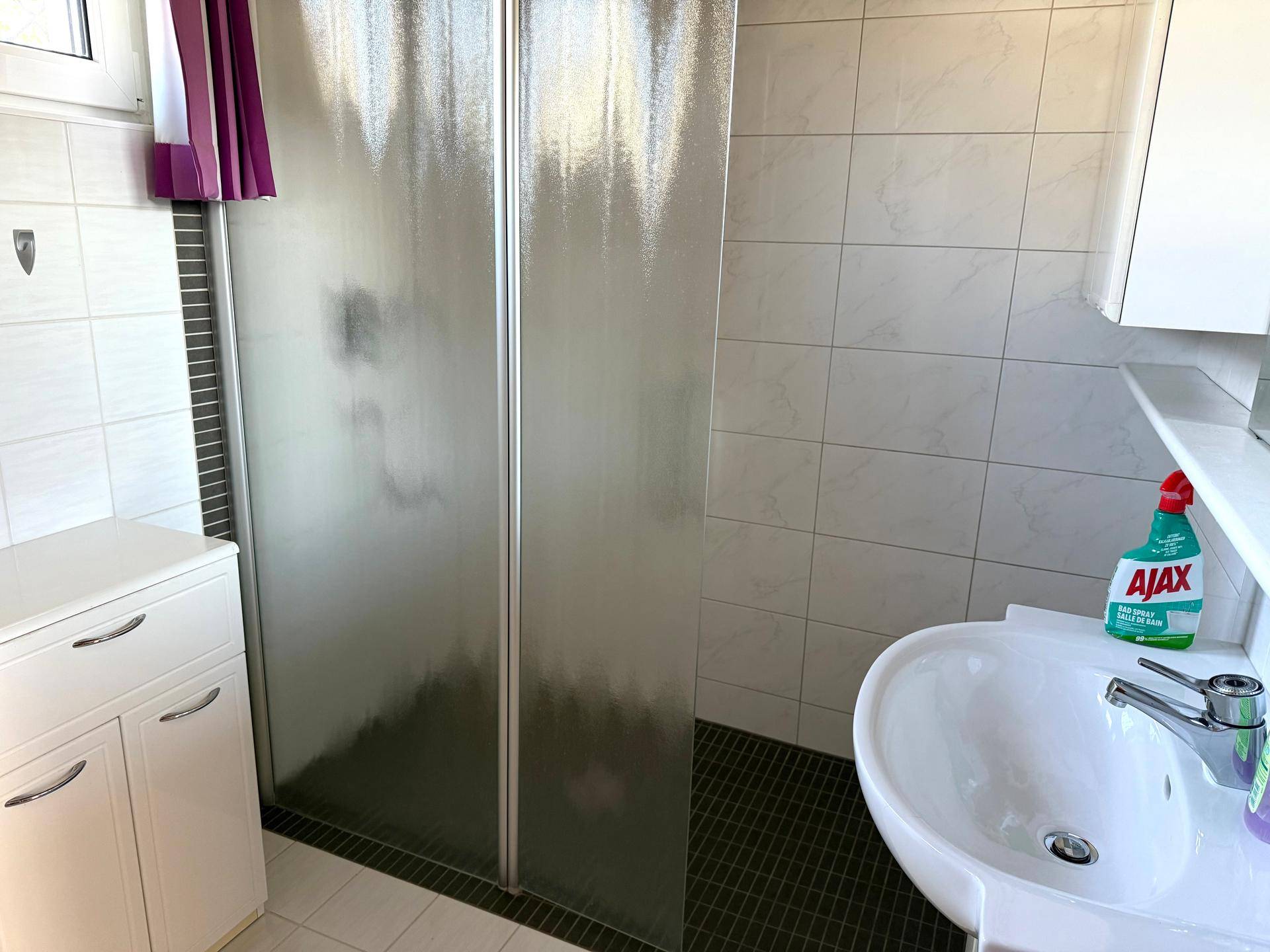 Badezimmer mit Duschkabine, Waschbecken und hellen Fliesen an Wänden und Boden.