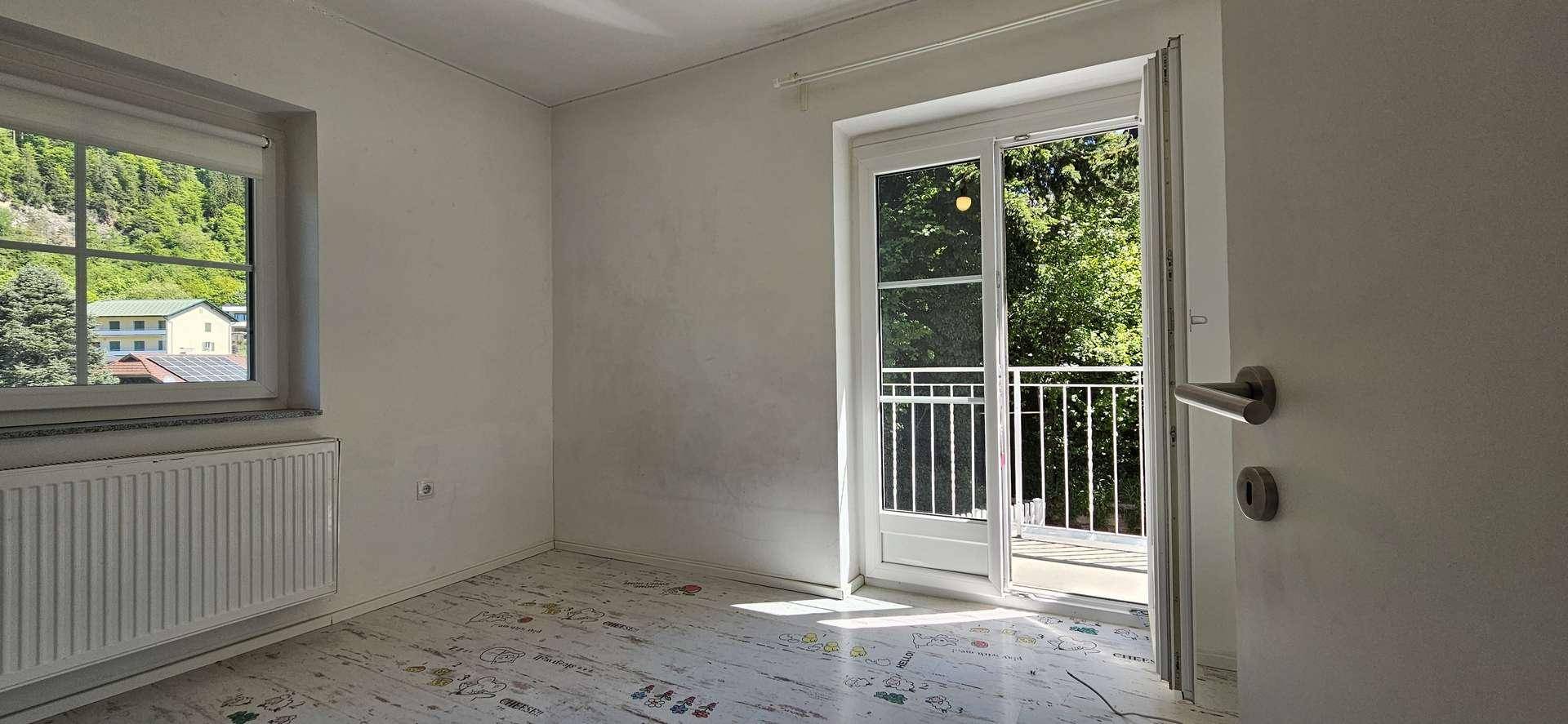 Helles Schlafzimmer mit gemustertem Boden, weißen Wänden und Balkonzugang, umgeben von viel Grün.