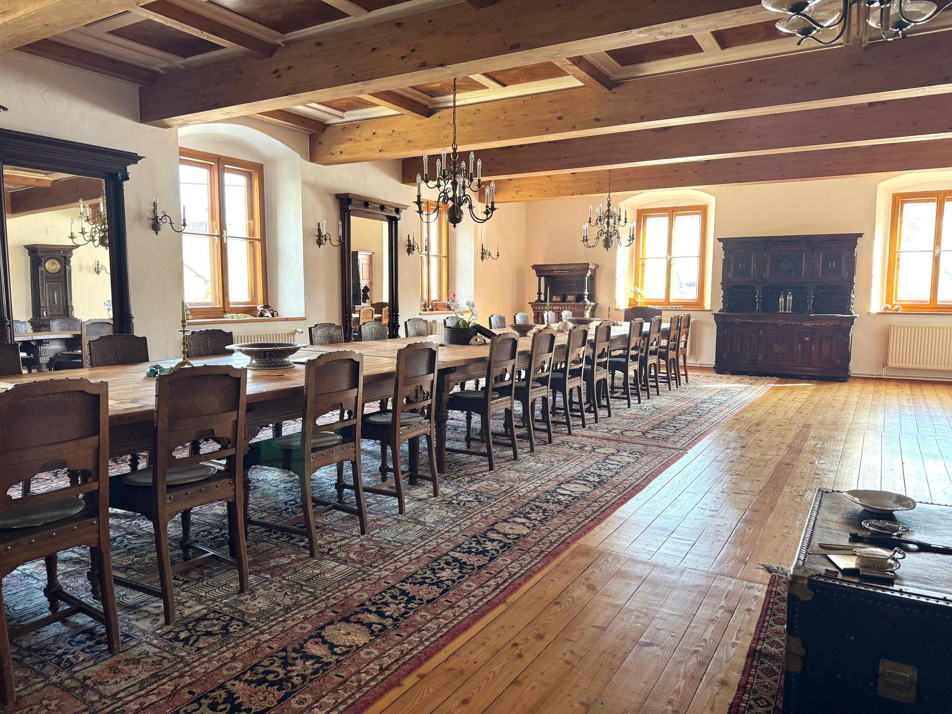 Weitläufiger Speisesaal mit langer Holztafel, Holzbalkendecke und vielen Fenstern.