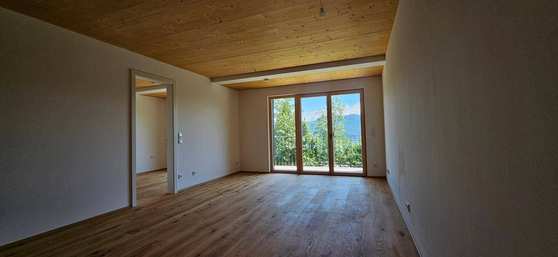 Heller Wohnraum mit Holzboden und Holzdecke, großen Fenstern und Zugang zum Balkon mit Bergblick.
