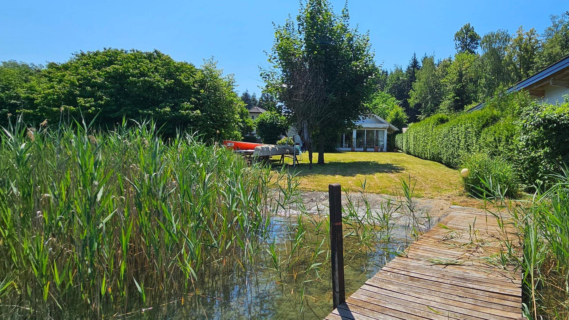 Holzsteg führt durch Schilf zum Seeufer, mit Blick auf das Haus und den Garten im Hintergrund.