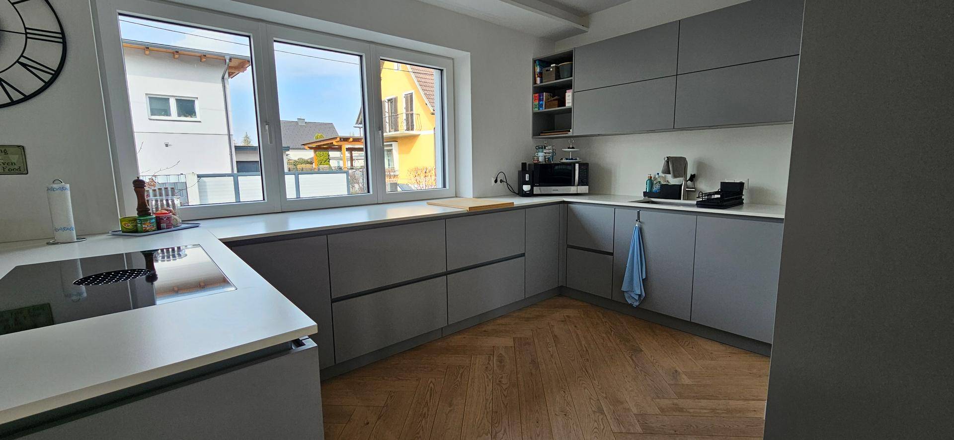 Helle Küche mit grauen Unterschränken, großem Fenster und Fischgrätparkettboden.