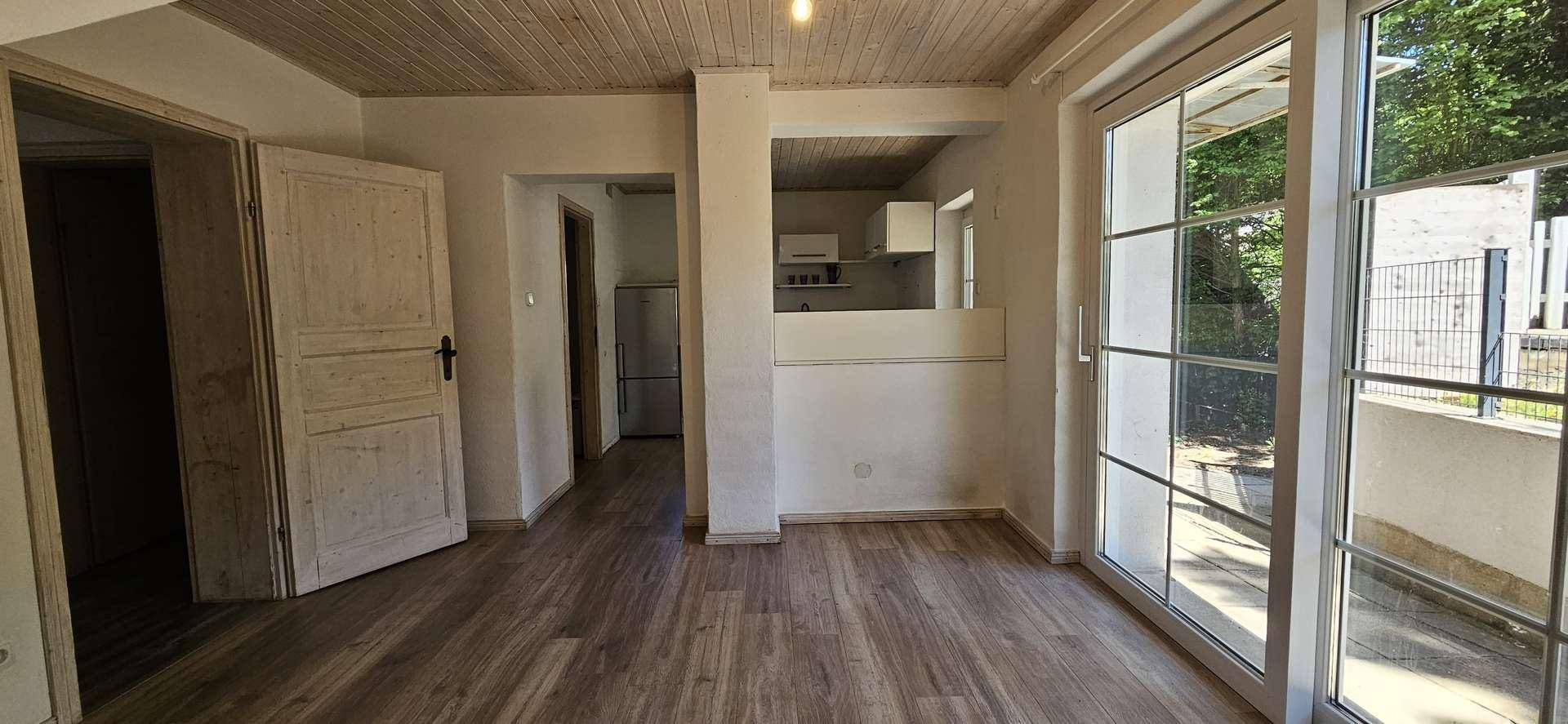 Heller Wohnbereich mit Holzdielen-Decke, Laminatboden und Blick auf eine halboffene Küche sowie Terrassenzugang.