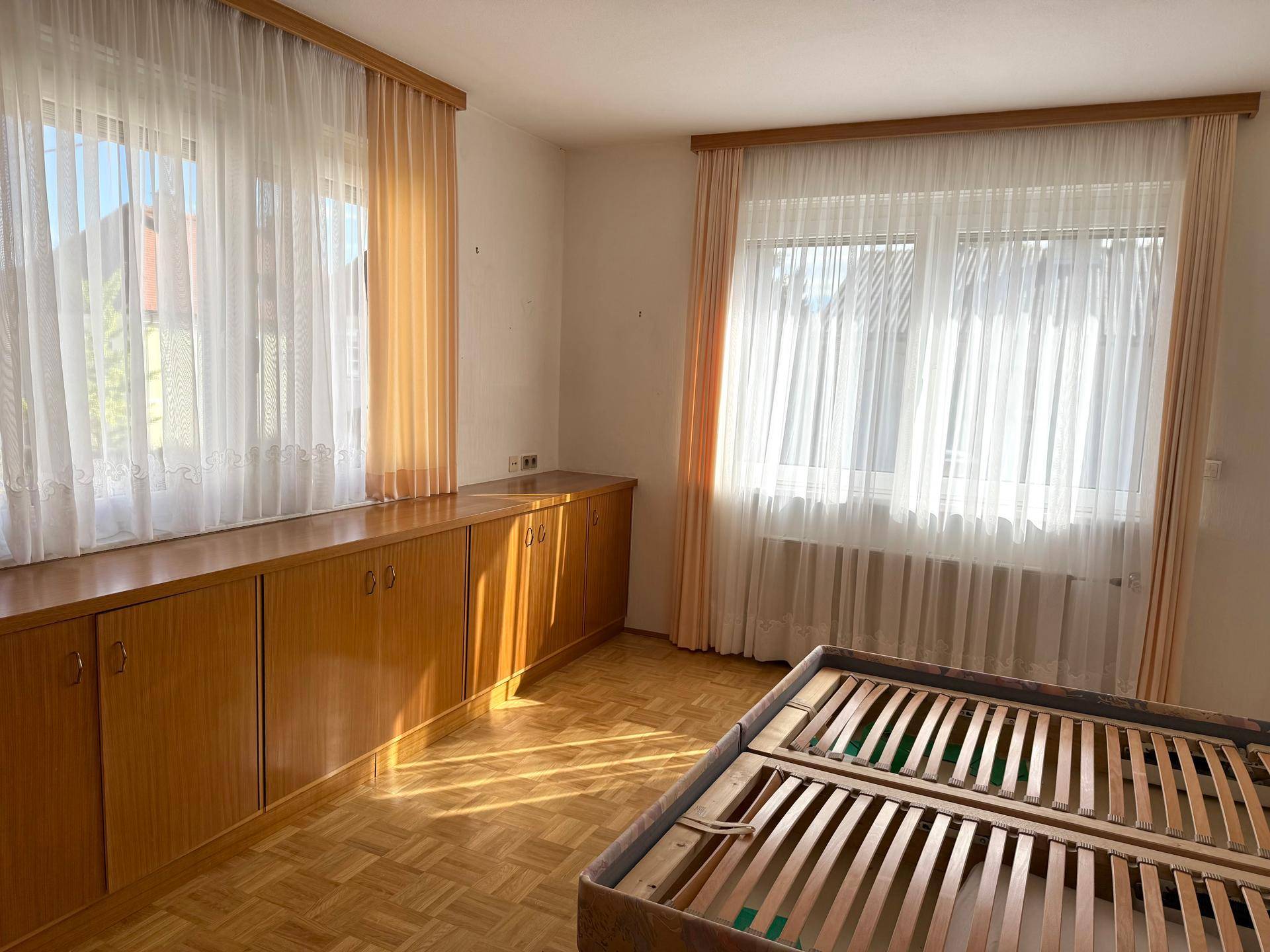 Schlafzimmer mit langen Einbauschränken aus Holz, Parkettboden und zwei großen Fenstern.