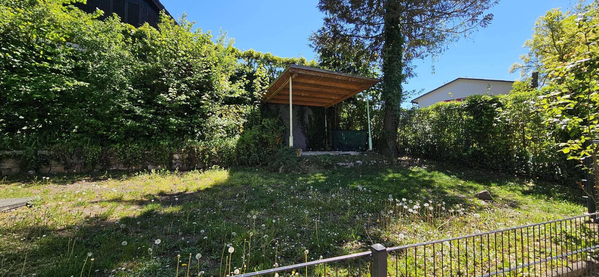 Gartenbereich mit einer kleinen Holzüberdachung, Rasenfläche und üppiger Bepflanzung im Hintergrund.