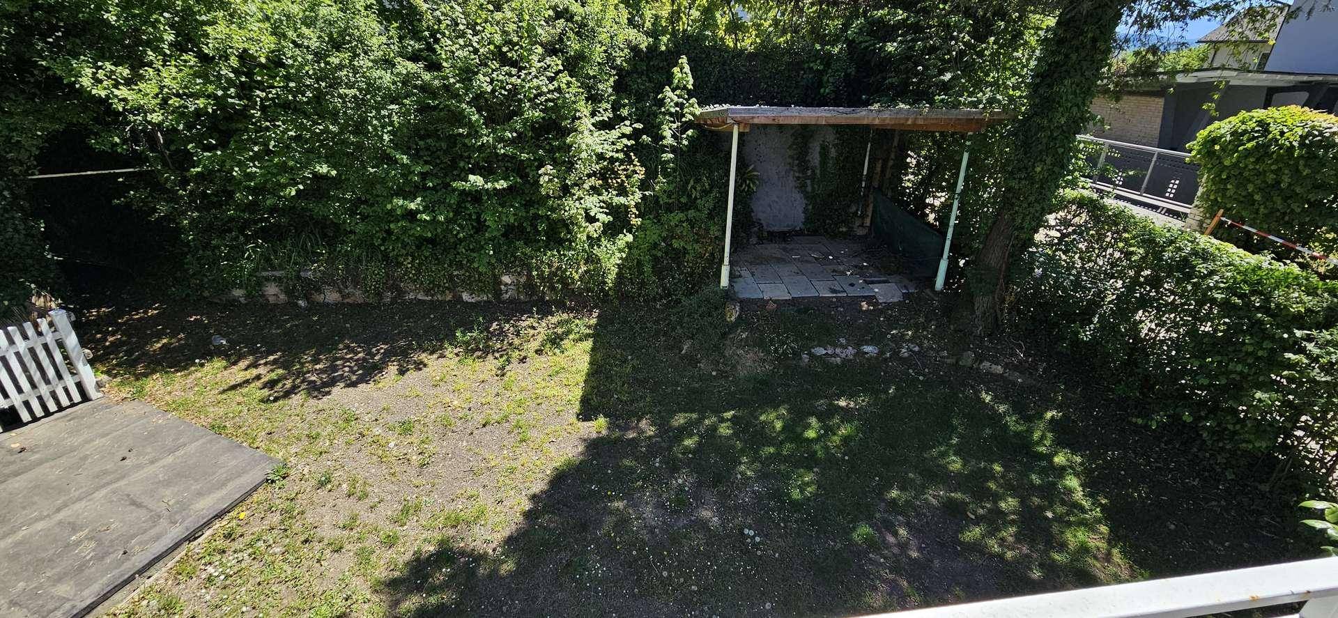 Blick vom Balkon auf einen grünen Gartenbereich mit einer überdachten Terrasse und dichter Bepflanzung.