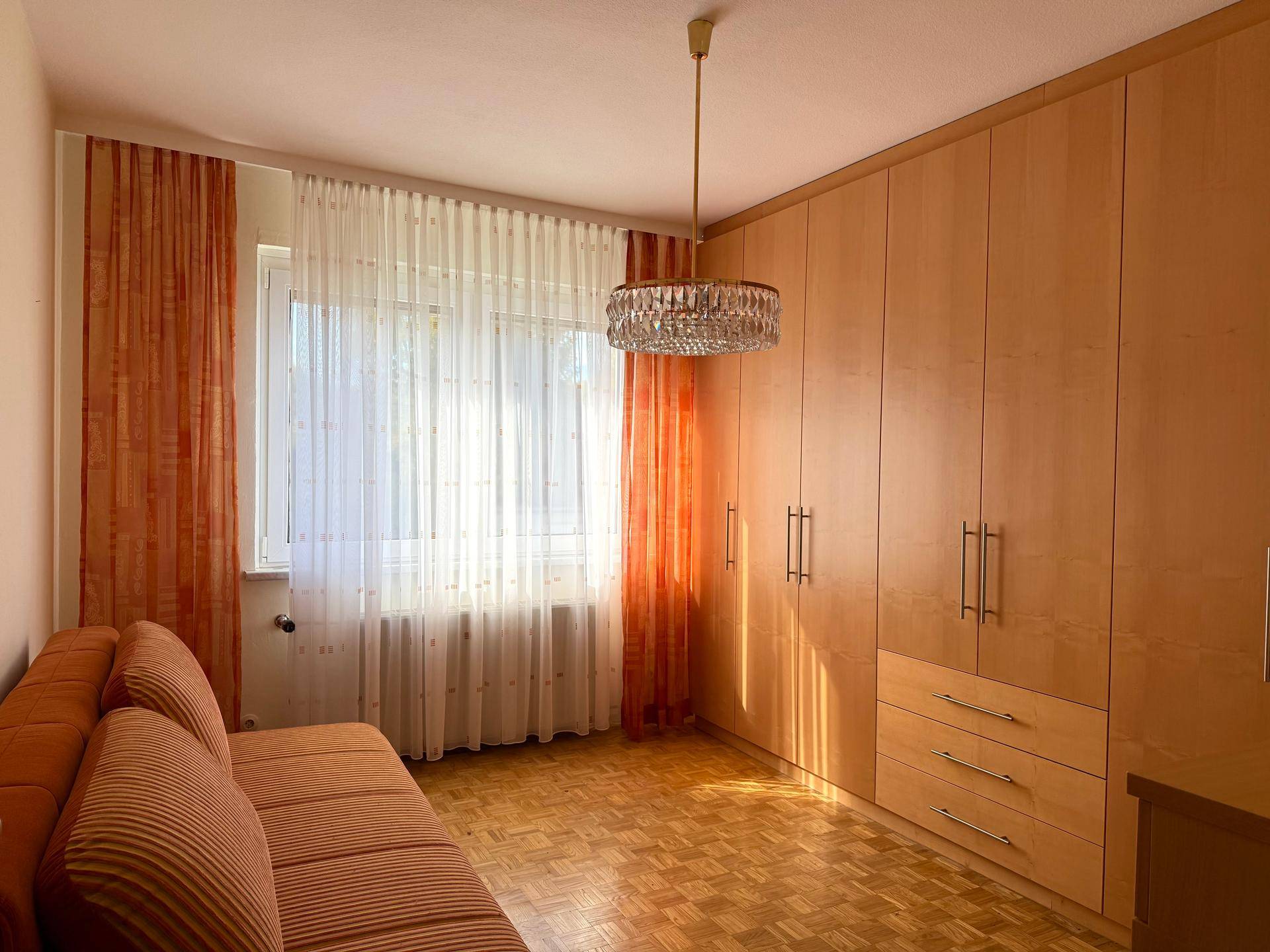 Schlafzimmer mit hellen Holzschränken, Parkettboden und viel Tageslicht durch das Fenster.